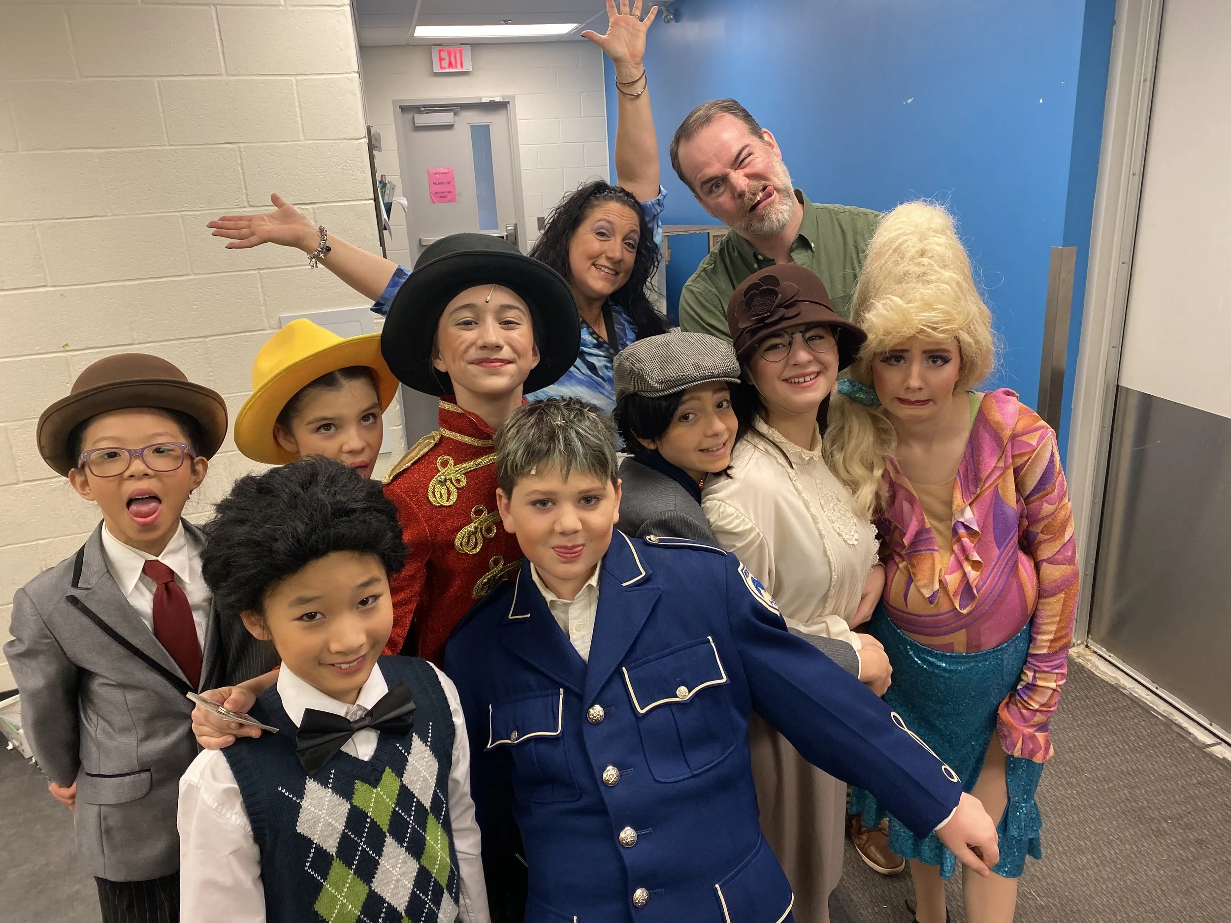 A group of children and adults dressed in costumes posing together in a hallway. The children are wearing various costumes, including a police uniform, a historical military outfit, and a fairy tale character. The adults are also in costumes, with on