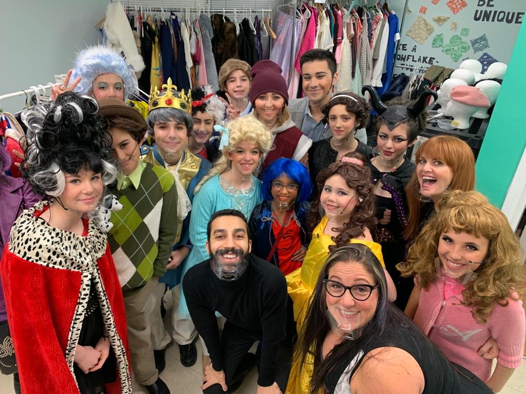Group of children and adults dressed in costumes, some with horns, wigs, or crowns, in a room with costumes and accessories hanging behind them.