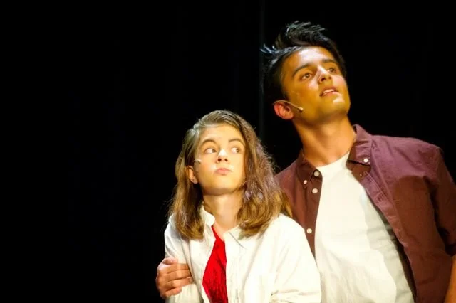 Two young performers, a girl and a boy, on stage during a theatrical performance, with the girl looking slightly concerned and the boy gazing upward with a confident expression.