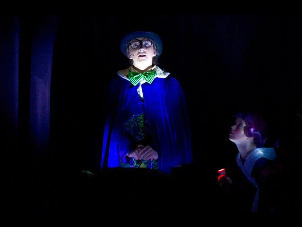 A person dressed as a wizard or magician with glasses and a blue robe, holding a book, standing in a dark environment with a spotlight; another person with purple hair looking up at the person in costume.
