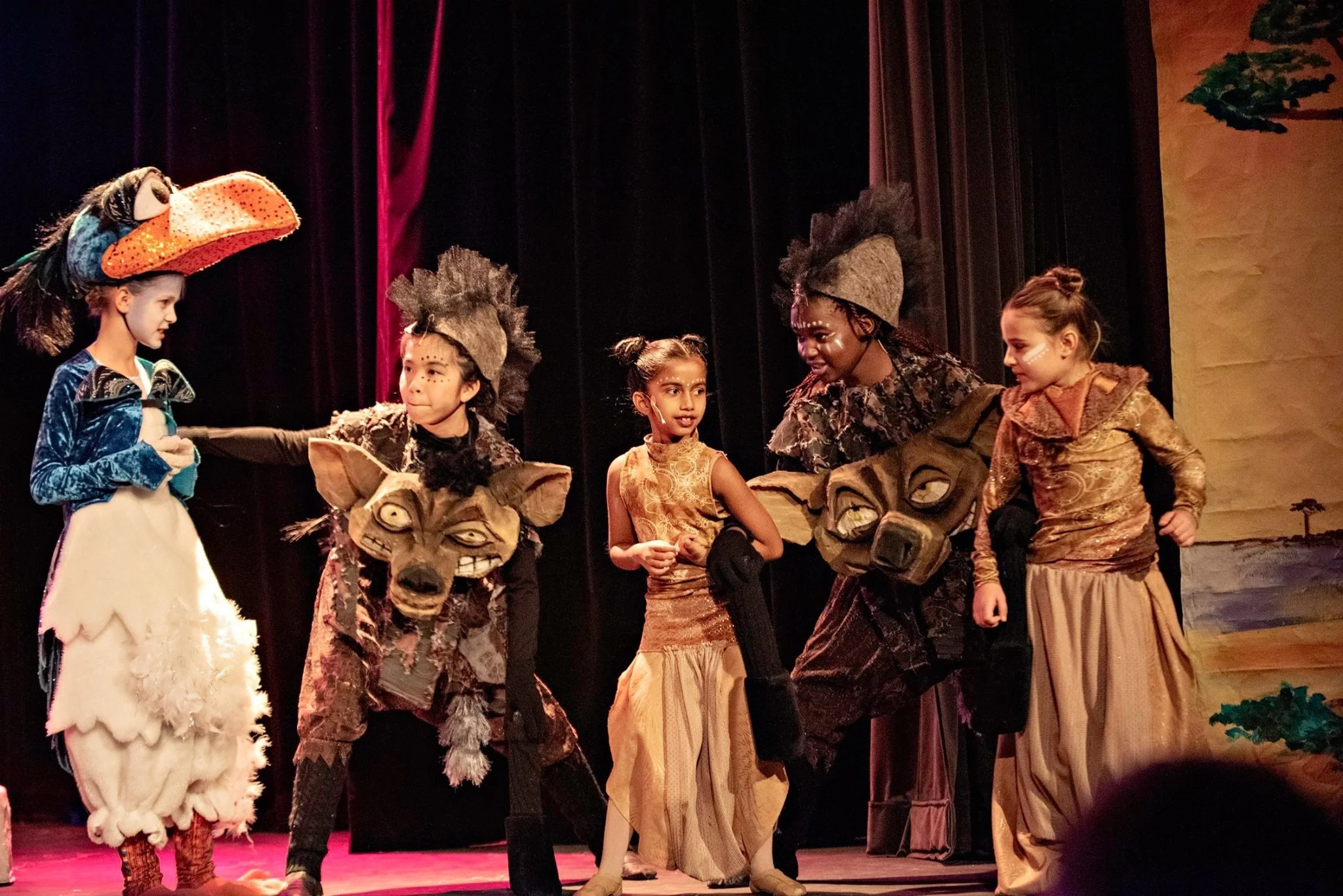 Children in costume performing on stage in a play or musical, with a black curtain and stage decorations in the background.