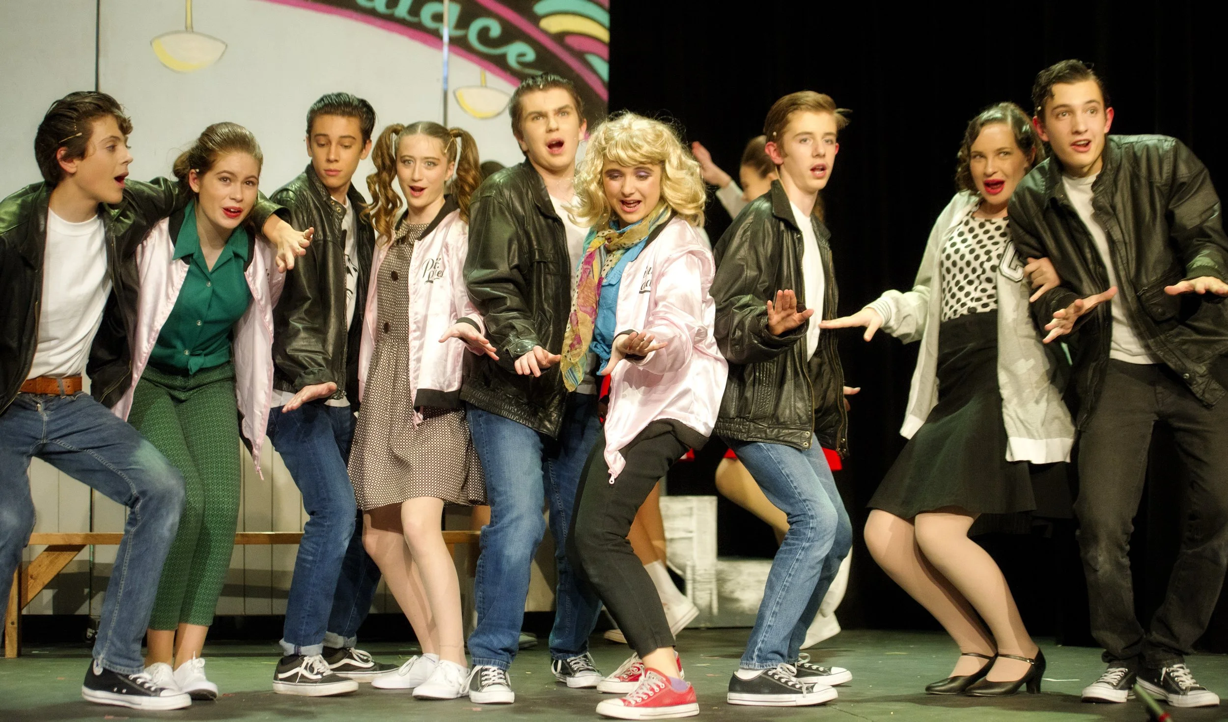 A group of young people performing on stage, dressed in 1950s style outfits. They appear to be singing or acting, with some expressive gestures and facial expressions. The background shows a colorful sign, indicating a theatrical or musical performan