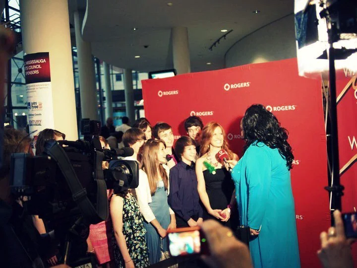 A group of children and an adult woman with curly black hair in an interview or photoshoot setting, in front of a red Rogers backdrop, with cameras and filming equipment present.