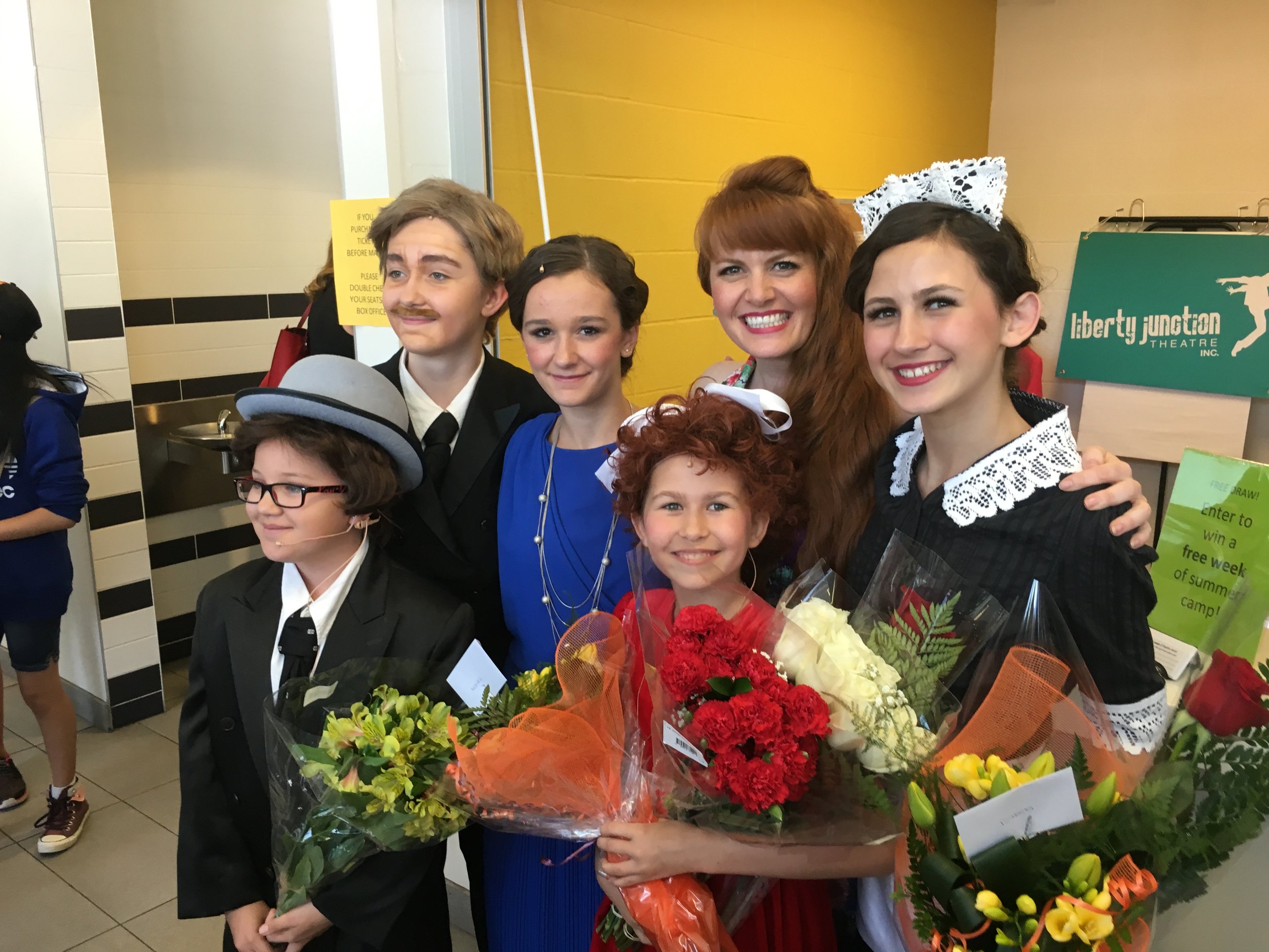 A group of children and an adult woman smiling, with the woman holding bouquet of flowers, standing near a yellow wall in an indoor setting.