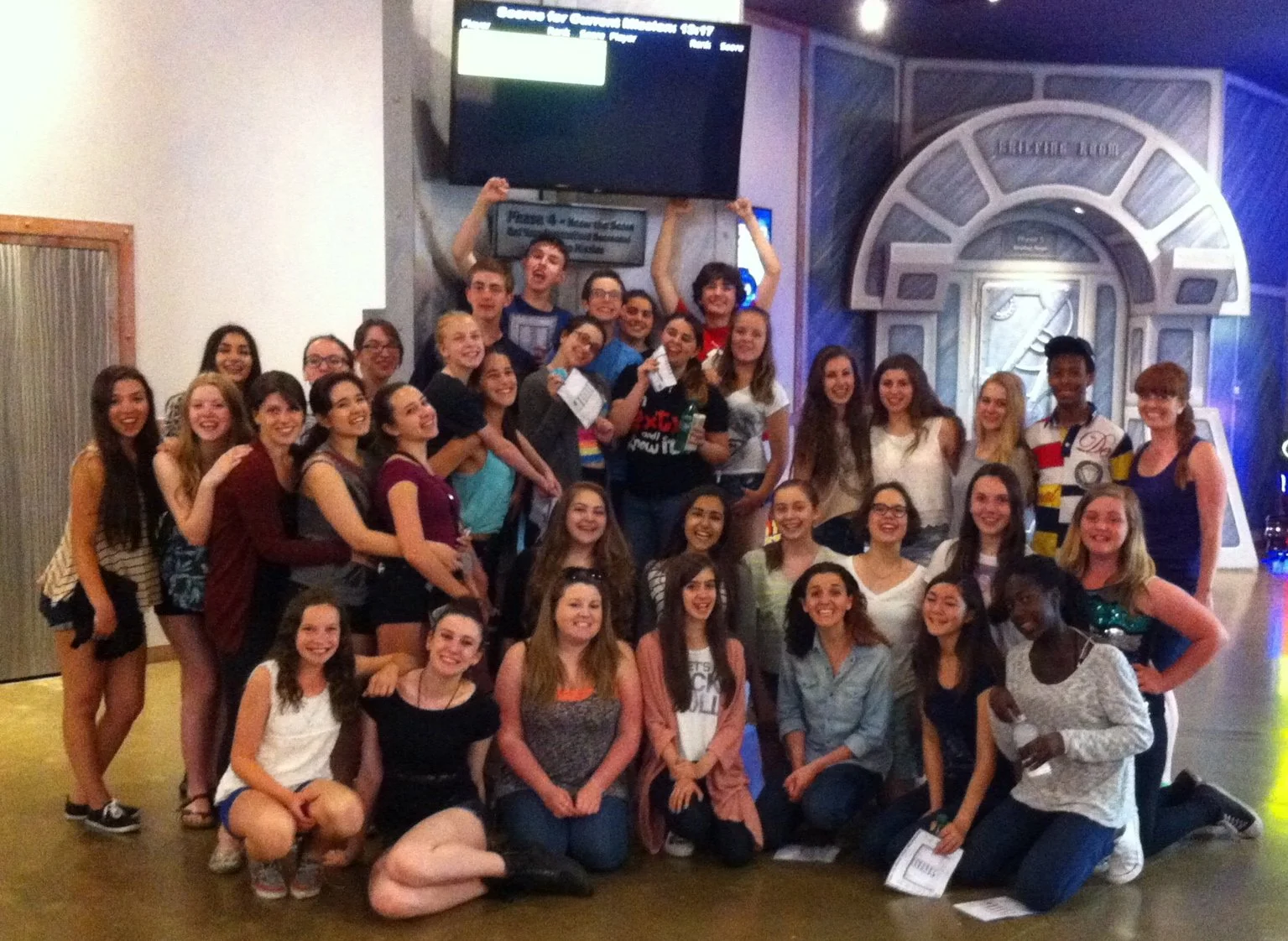 A large group of young people smiling and posing for a group photo indoors, with some holding papers and drinks, in front of a decorative background and a television screen.