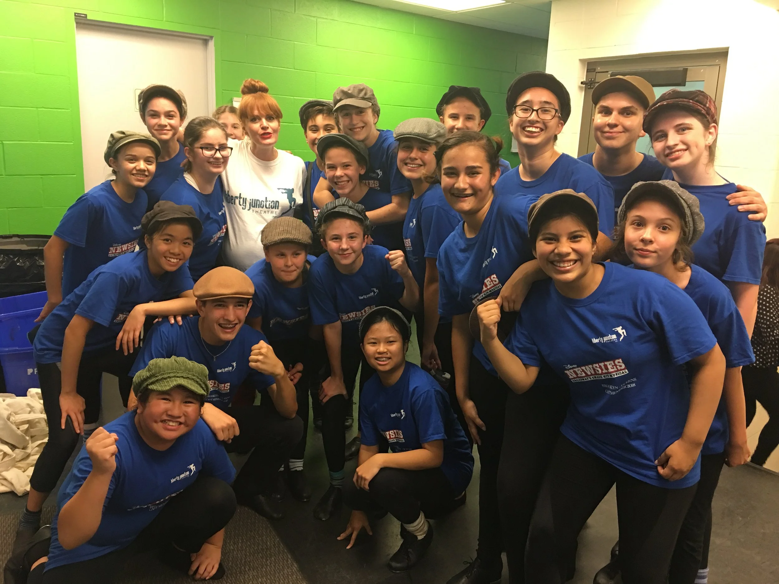 A group of young children wearing blue T-shirts and vintage-style hats, smiling and posing together in an indoor setting with a green wall in the background.