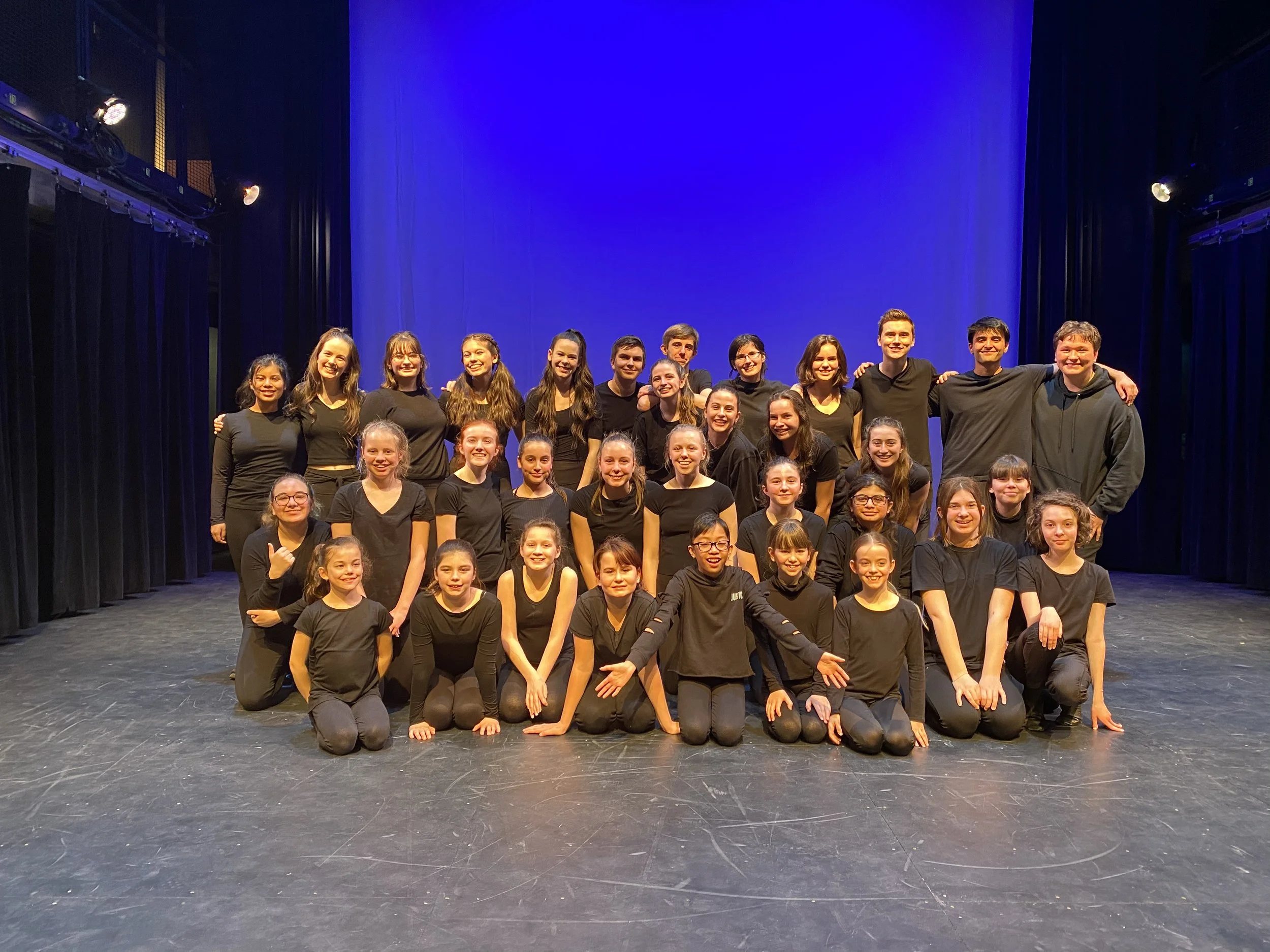 A group of young people, wearing black outfits, are smiling and posing for a group photo on a stage with a blue background.