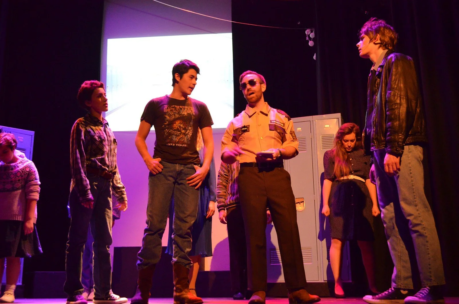 A group of teenagers and a police officer on stage, seemingly in a school play or drama. The police officer is speaking to one of the teens, with some of the teens looking on and others appearing upset or contemplative. Lockers are in the background.