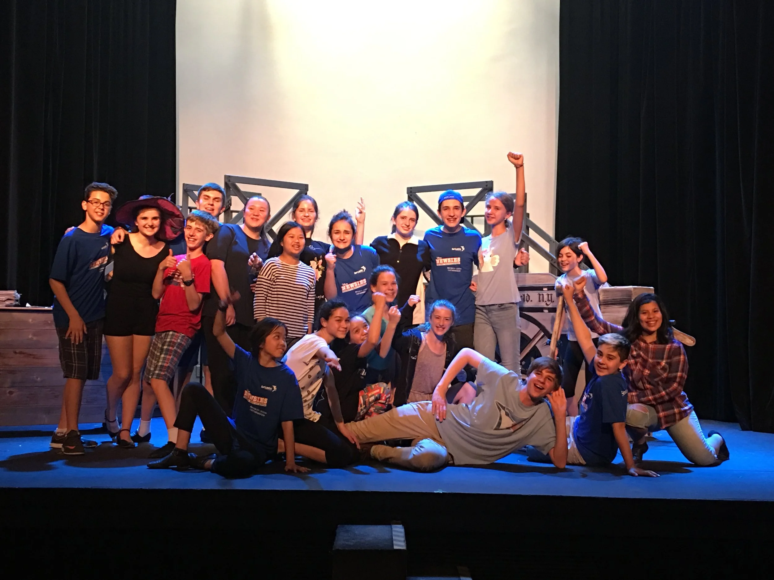 A group of children and teenagers on stage, many smiling, some making gestures, with festival or school shirts, behind a stage set that resembles a historical or theater scene, with stairs and a nameplate indicating New York, N.Y.