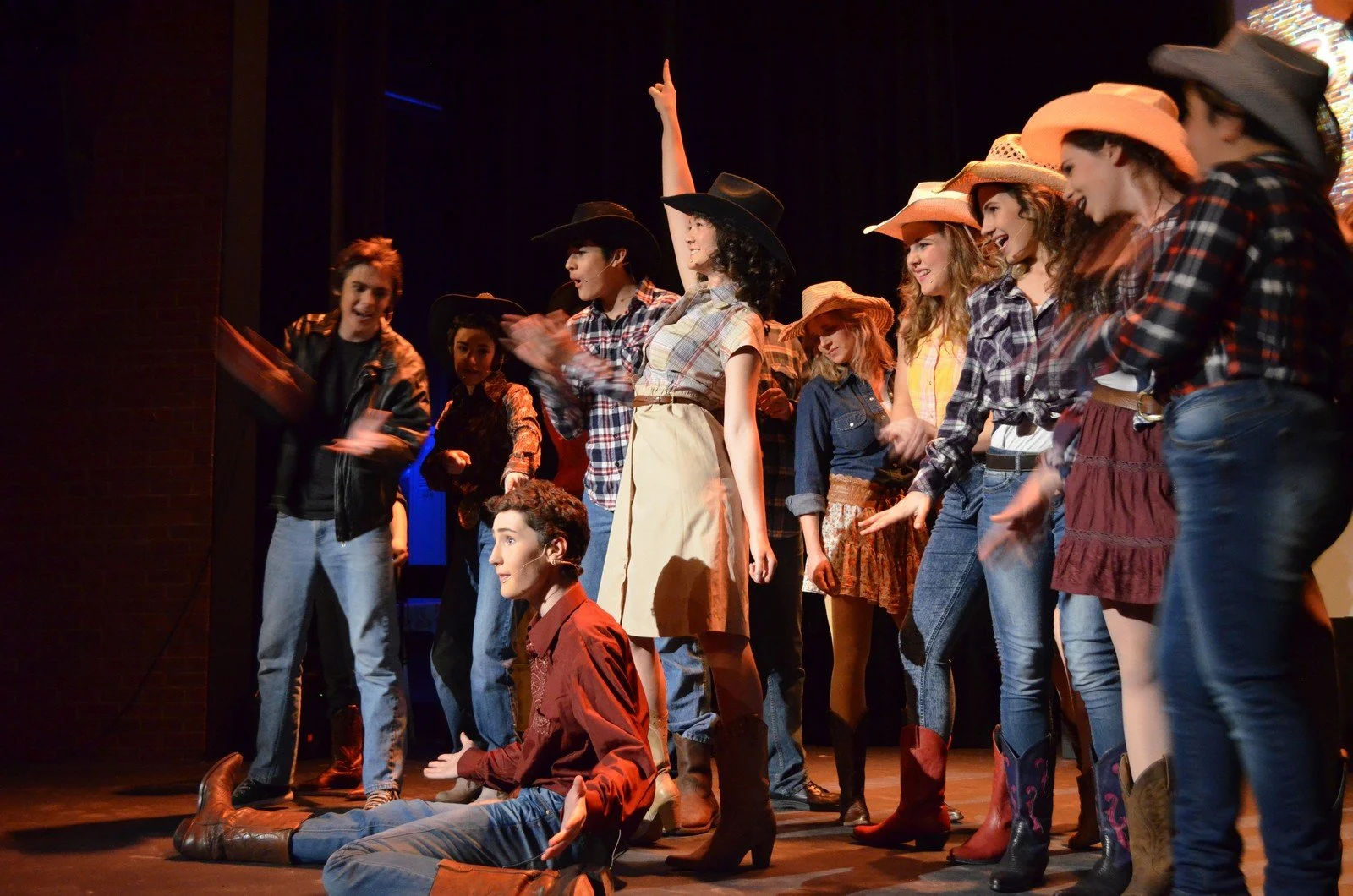 Theater cast in cowboy costumes on stage, one person lying on floor, others celebrating, in a Western-themed play.