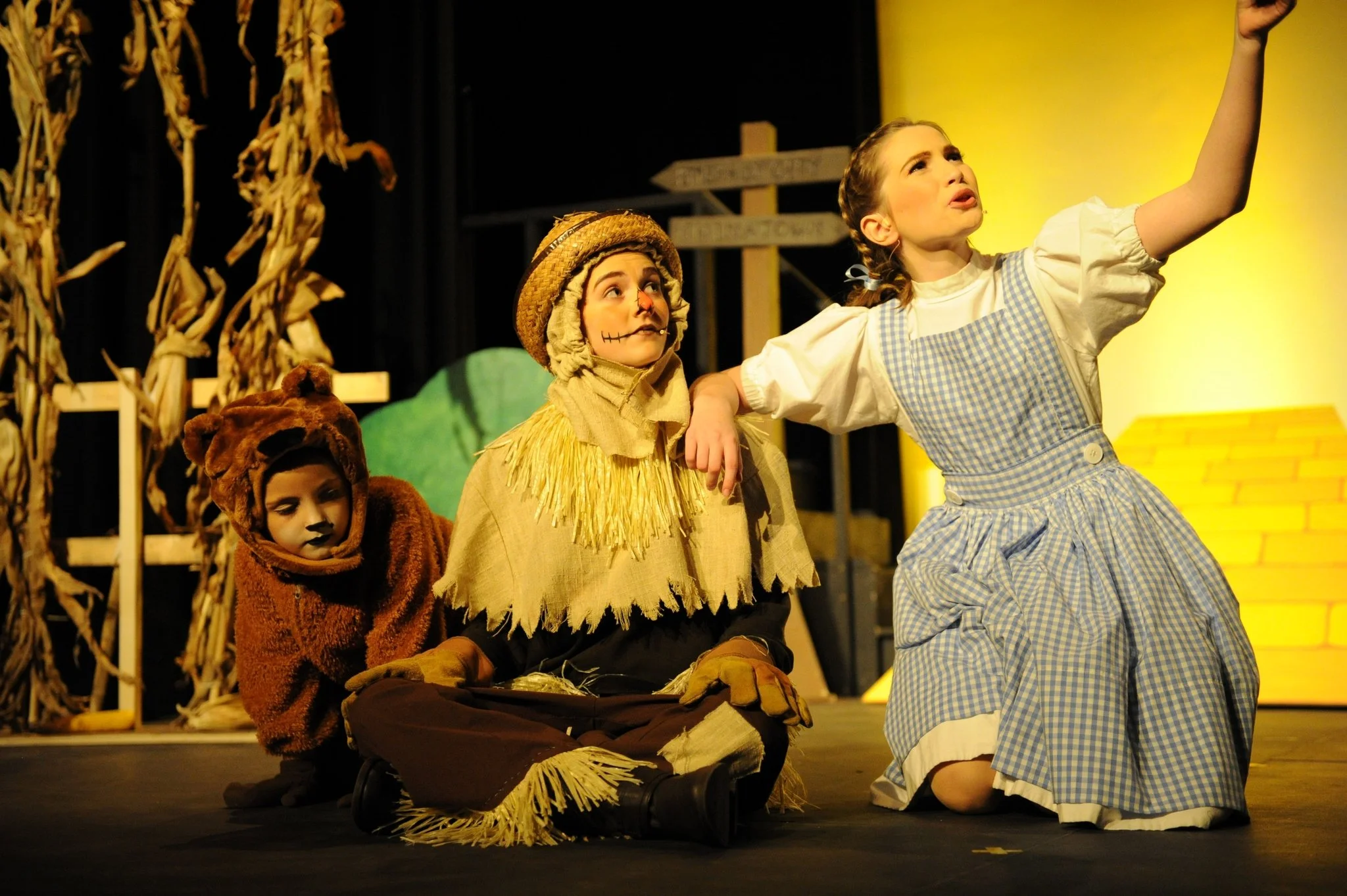 Children dressed as characters from The Wizard of Oz on stage, with one girl as Dorothy and others as scarecrow, lion, and cowardly lion, performing in a theatrical production.