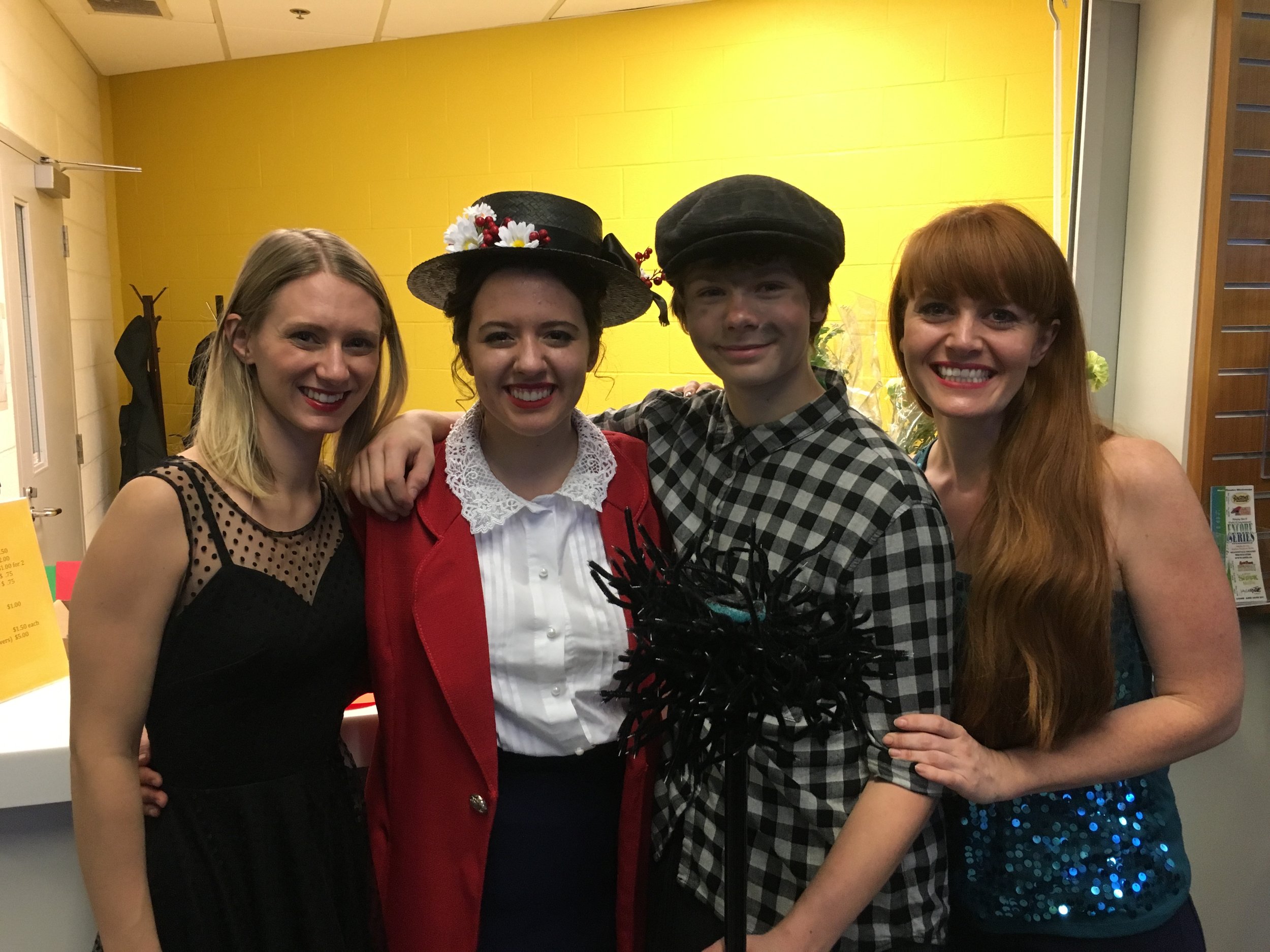 Four people smiling and standing close together indoors, with bright yellow wall, dressed in vintage-style clothing and hats, holding a black decorative object.