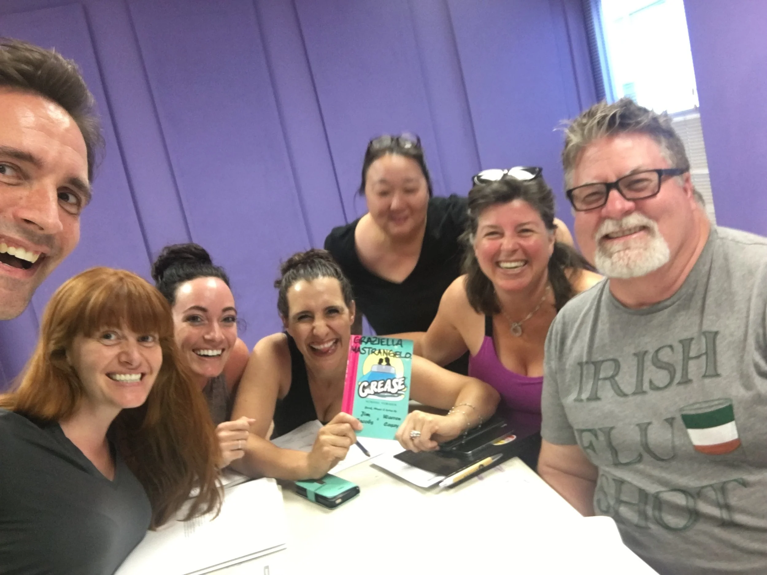 Group of smiling people gathered around a table, holding a colorful card that says 'GREASE'. Purple wall in the background, some people wearing glasses, some with hair tied back.