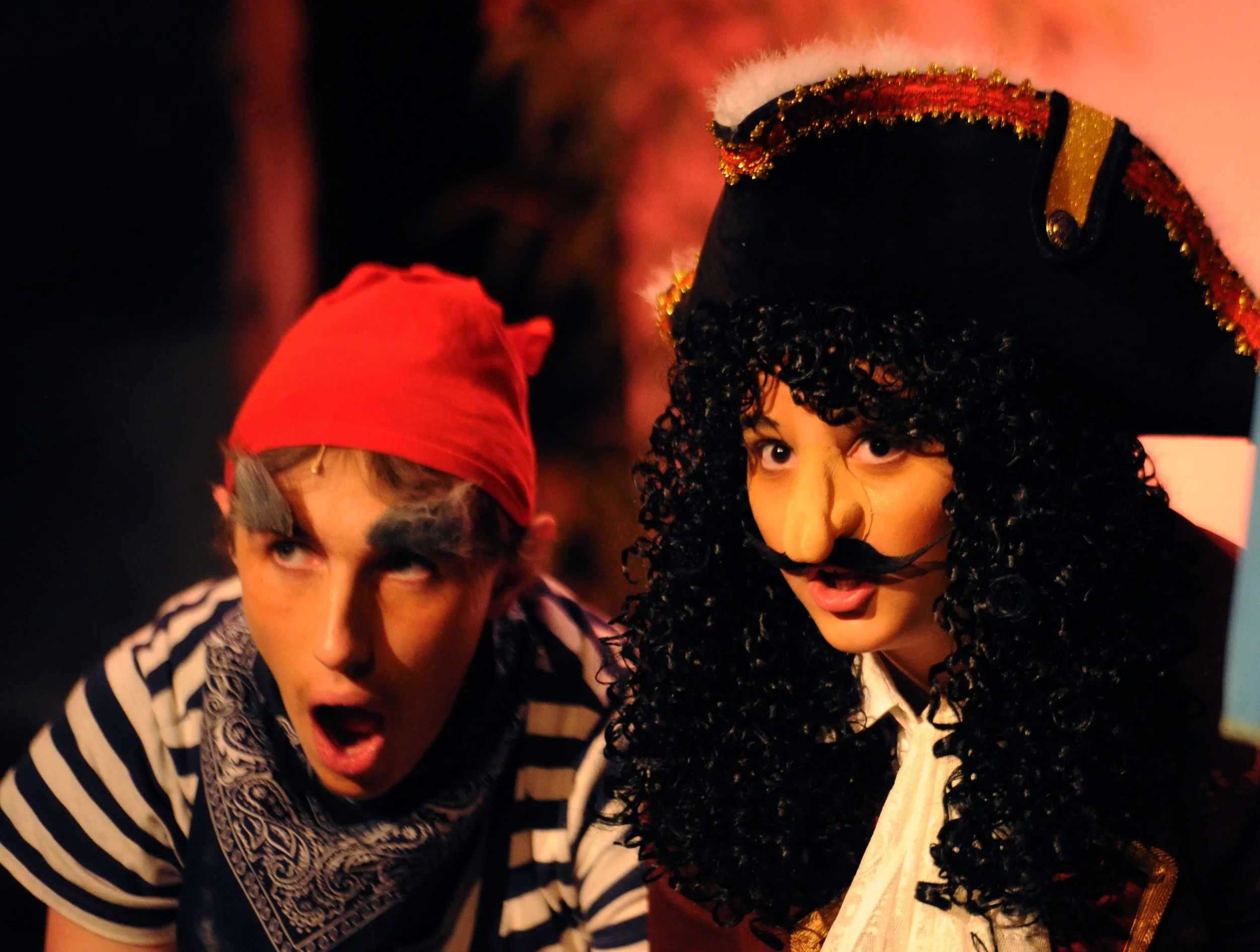 Two people dressed in costumes with a black background. The person on the left is wearing a red hat and a striped shirt, with an open mouth and eyes looking up. The person on the right is wearing a curly black wig, a colorful hat with gold trim, and 