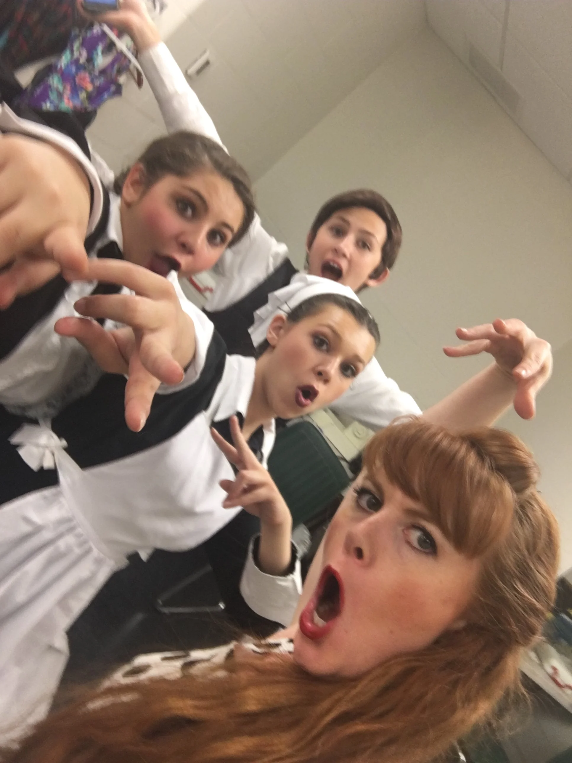 Four women in white and black uniforms making playful faces and gestures in a room with a plain ceiling and background.