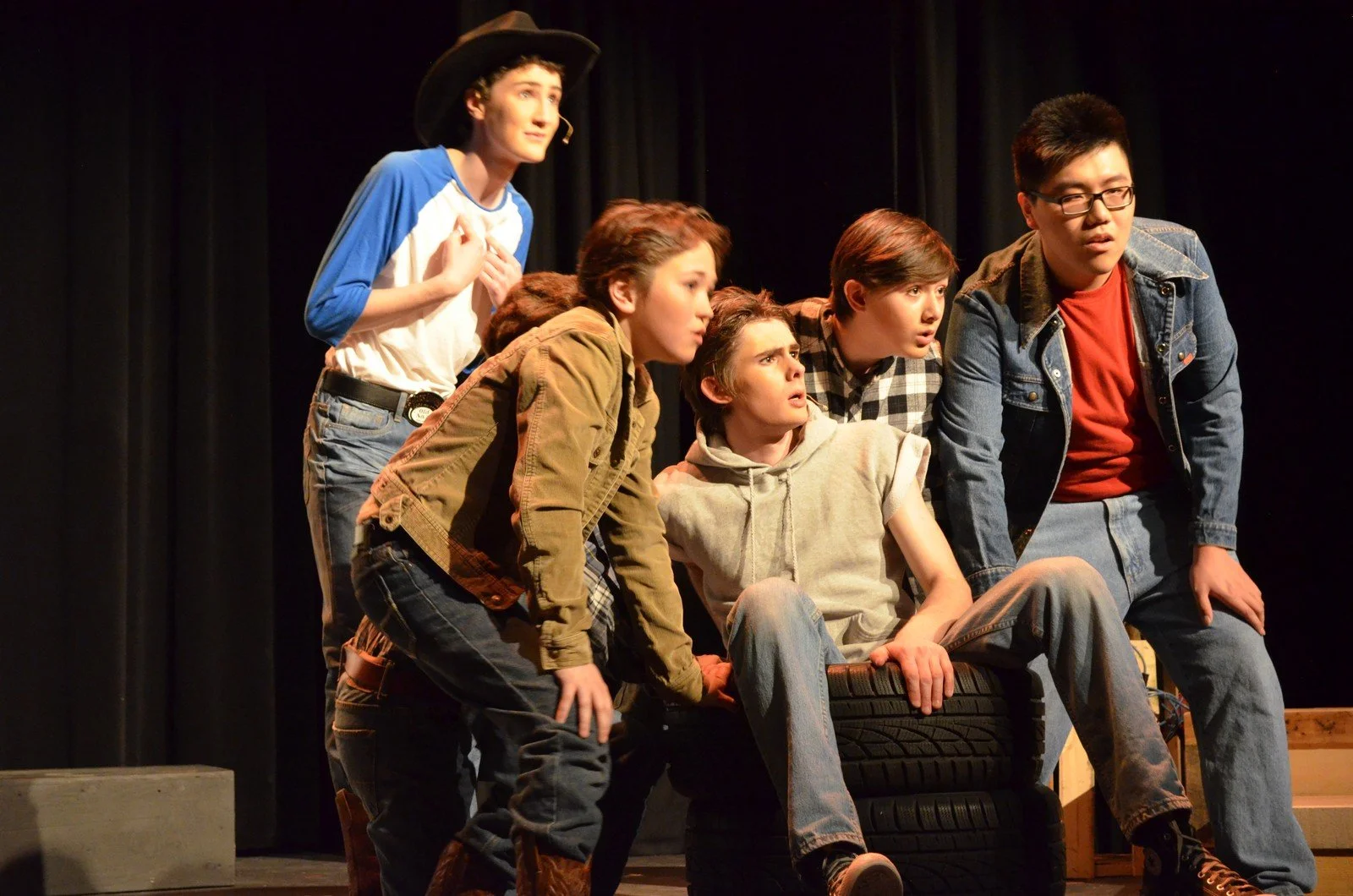 A group of six young actors performing on stage, with some leaning forward and others sitting on tires, with black curtains in the background.