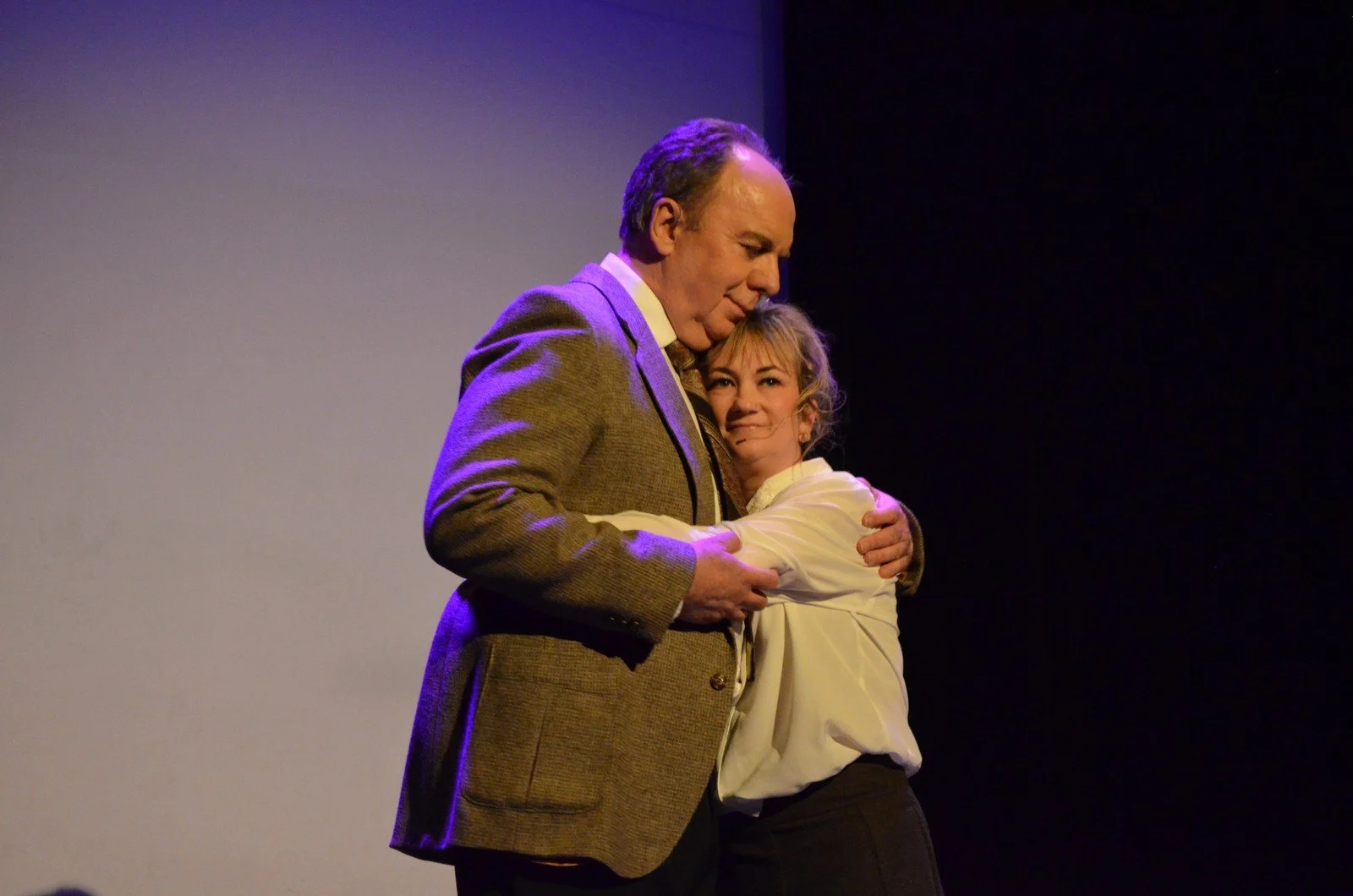 Two actors on stage, one man and one woman, hugging each other. The man is wearing a brown jacket and the woman has a white shirt. They appear emotional, with the woman looking at the camera and the man resting his head on her. The background is dark