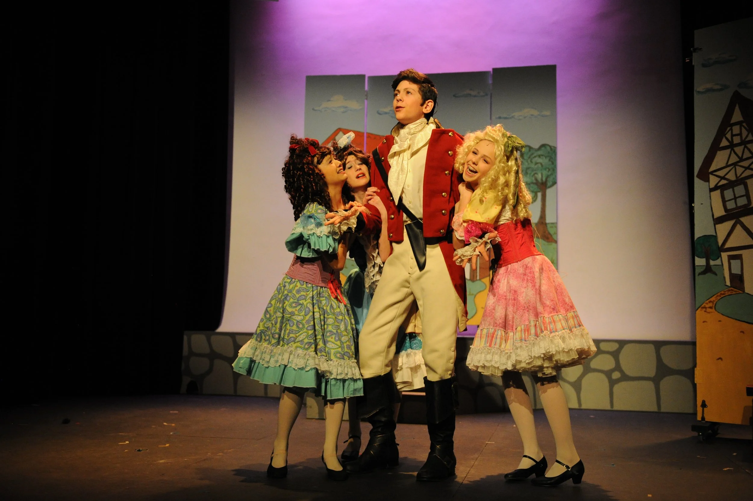 Stage performance with four actors in historical costumes. One male actor stands in the center wearing a red coat and beige trousers, looking upward. Three female actors surround him, wearing colorful, ruffled dresses and wigs, engaging with the male
