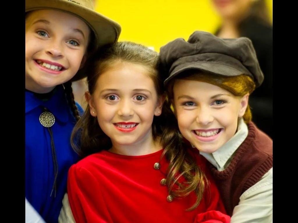 Three children dressed in vintage-style costumes, smiling and posing closely together in a cheerful setting.