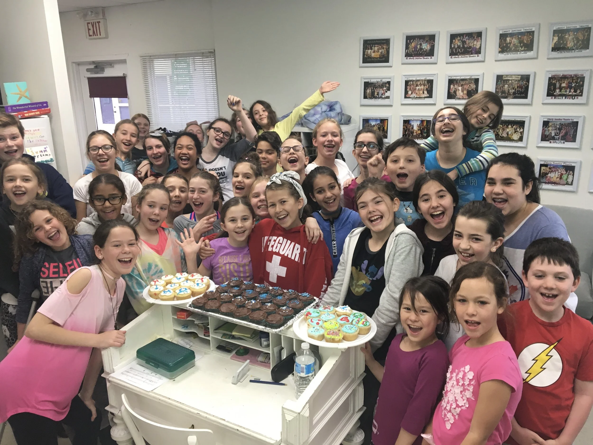 A group of children gathering around a table with cupcakes decorated with colorful icing and toppings, celebrating in a room with photos on the wall.