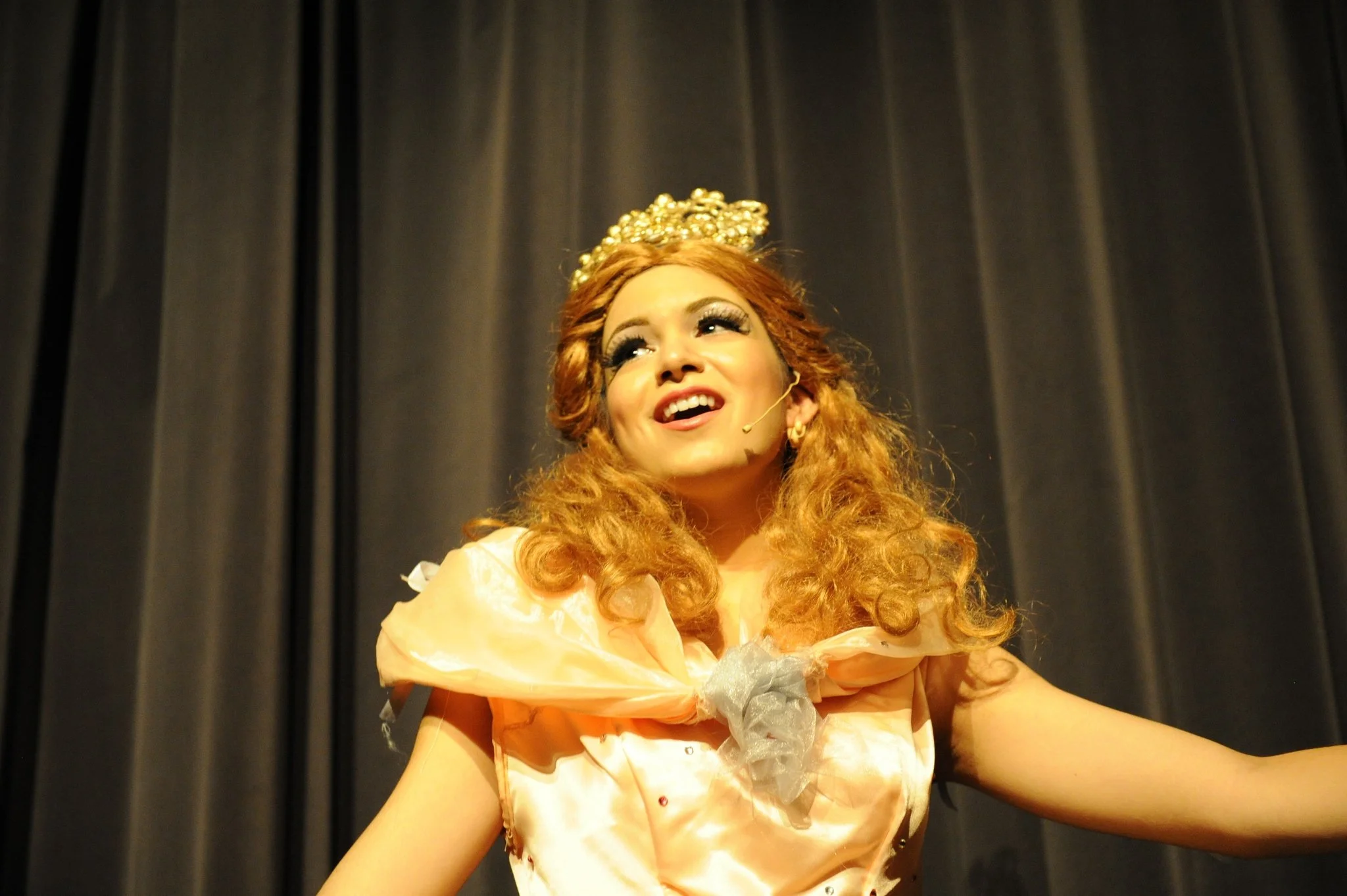 A woman dressed in a peach-colored gown with a crown or headpiece made of gold or shiny material, smiling and sitting against dark curtains.