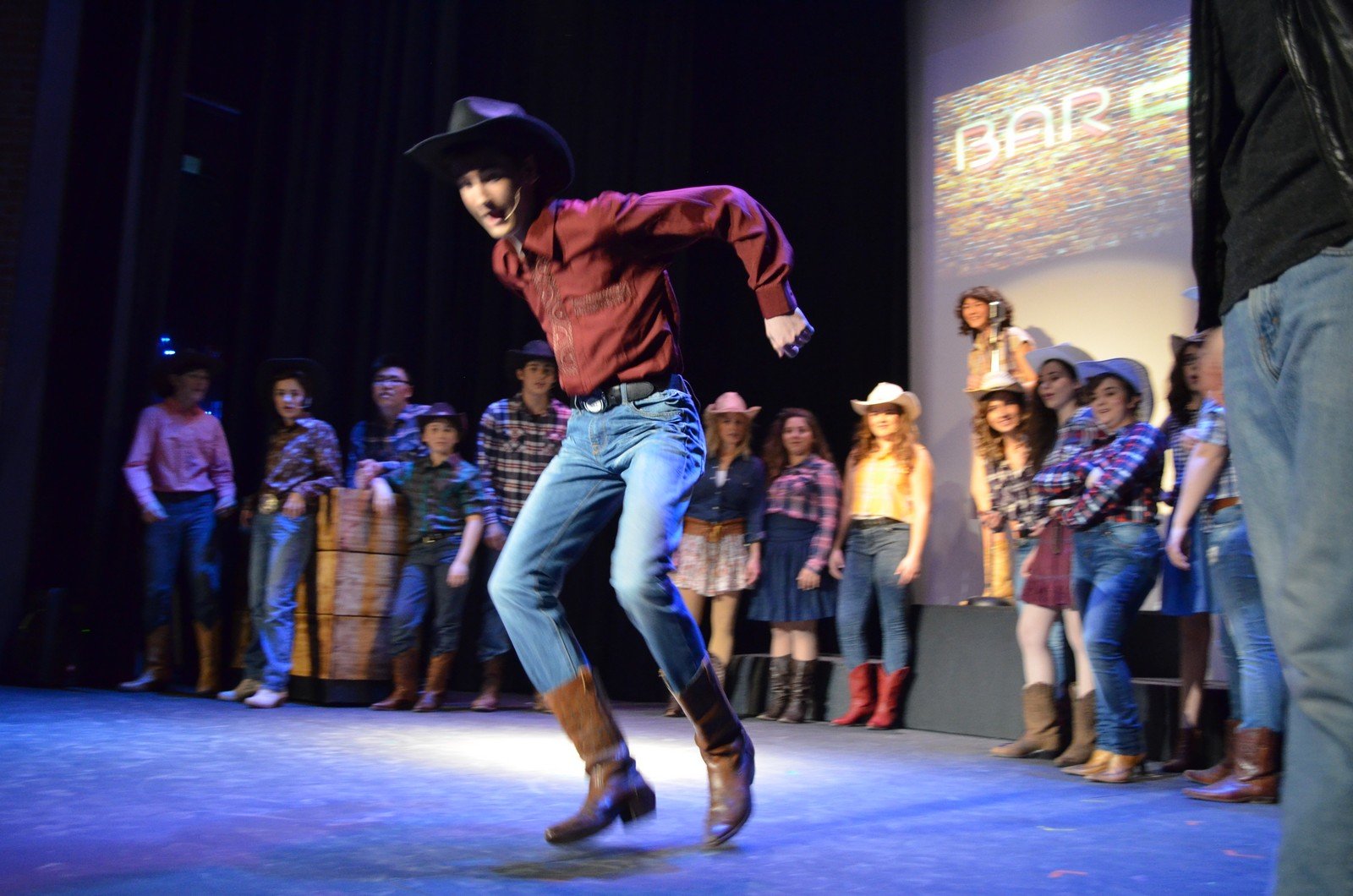 A young person in cowboy attire is dancing on stage with a group of children dressed in cowboy and cowgirl outfits watching from the background, in a setting resembling a country or western themed event.