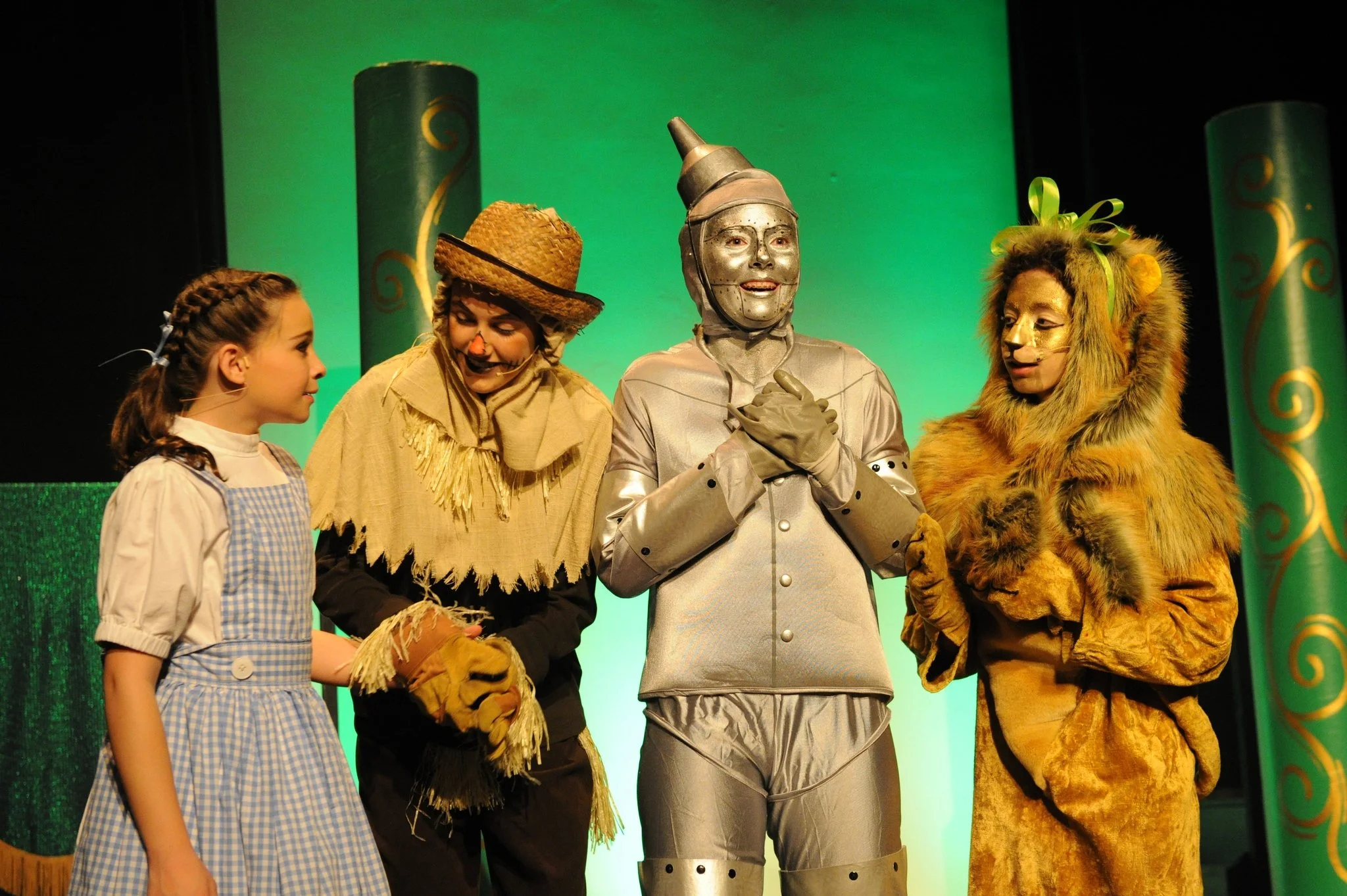 Four characters in costumes from The Wizard of Oz, including Dorothy, the Scarecrow, Tin Man, and Cowardly Lion, standing on stage.