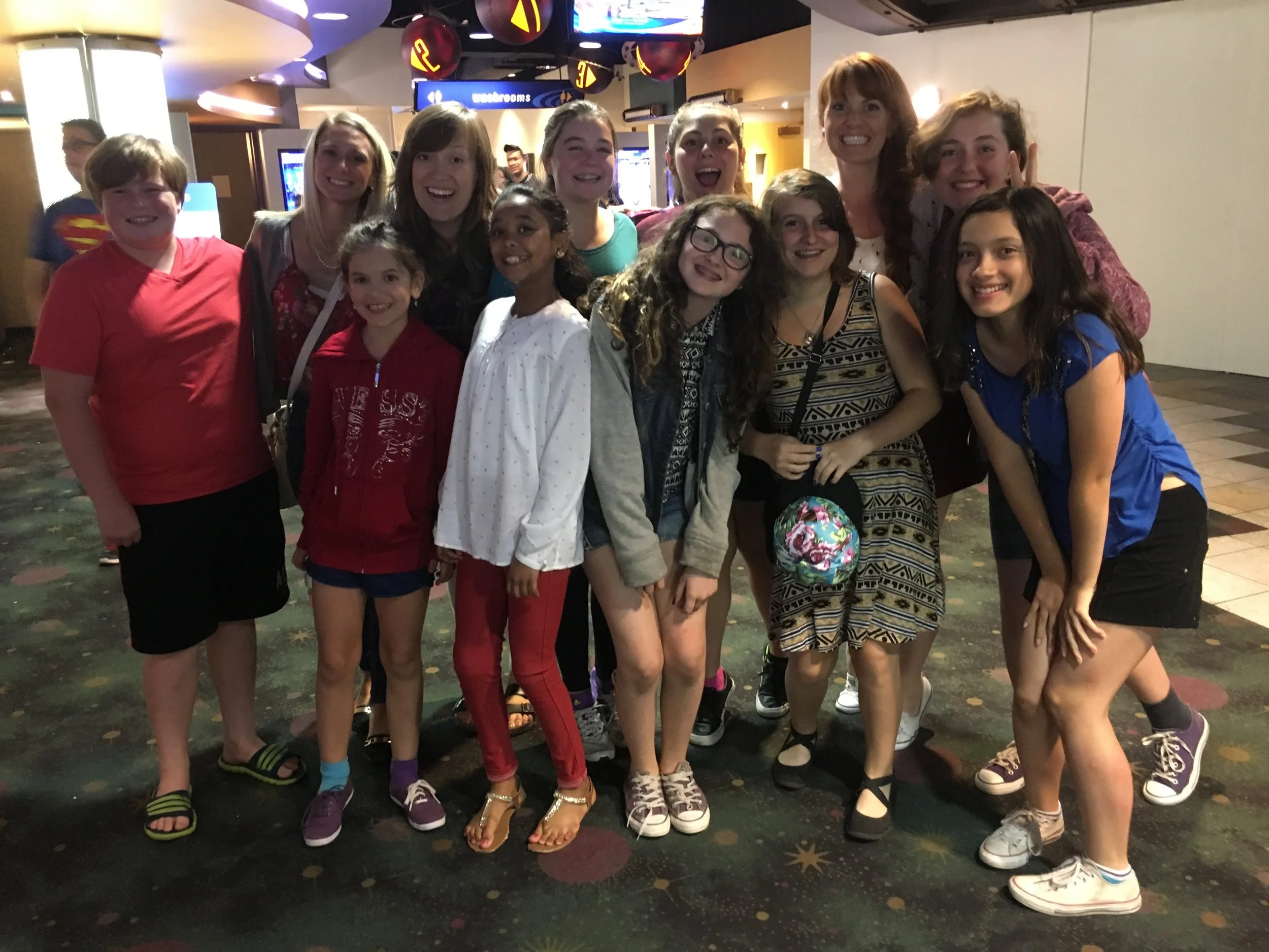 A group of young girls and women posing happily inside a colorful indoor entertainment venue, possibly a bowling alley, with bright lights and arcade games visible in the background.