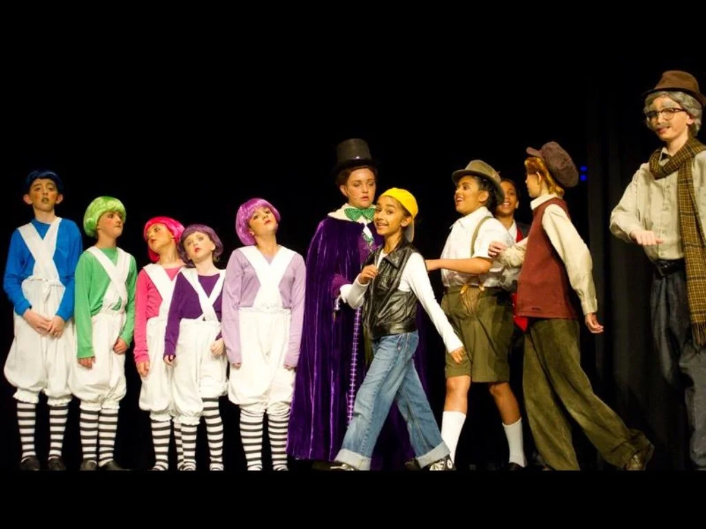 Children performing in a school play with colorful costumes resembling characters from a story, standing on stage against a black background.