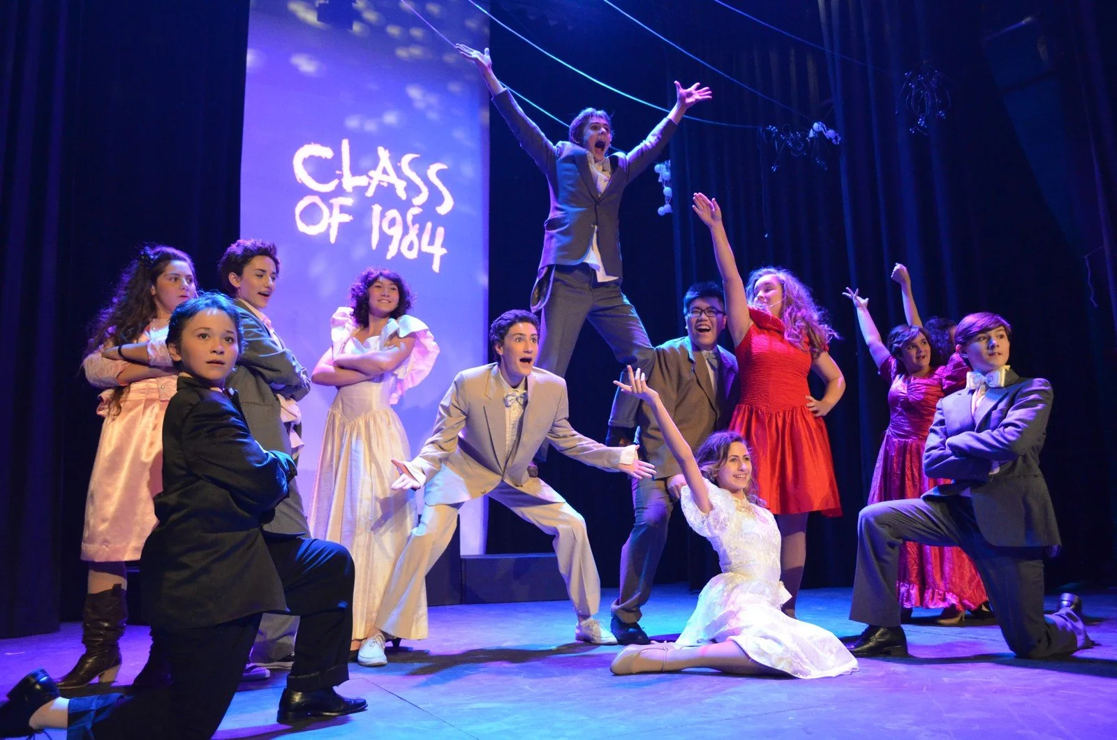 A theatrical performance of 'Class of 1984' features a young cast in vintage costumes, with some standing, some kneeling, and one person in the center jumping with arms raised against a blue backdrop displaying the show's title.