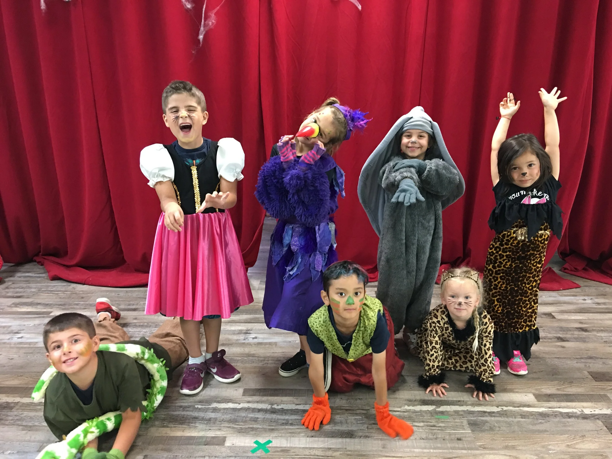 Children dressed in colorful costumes, some with face paint, standing and kneeling on a wooden floor in front of a red curtain, during a costume or school event.