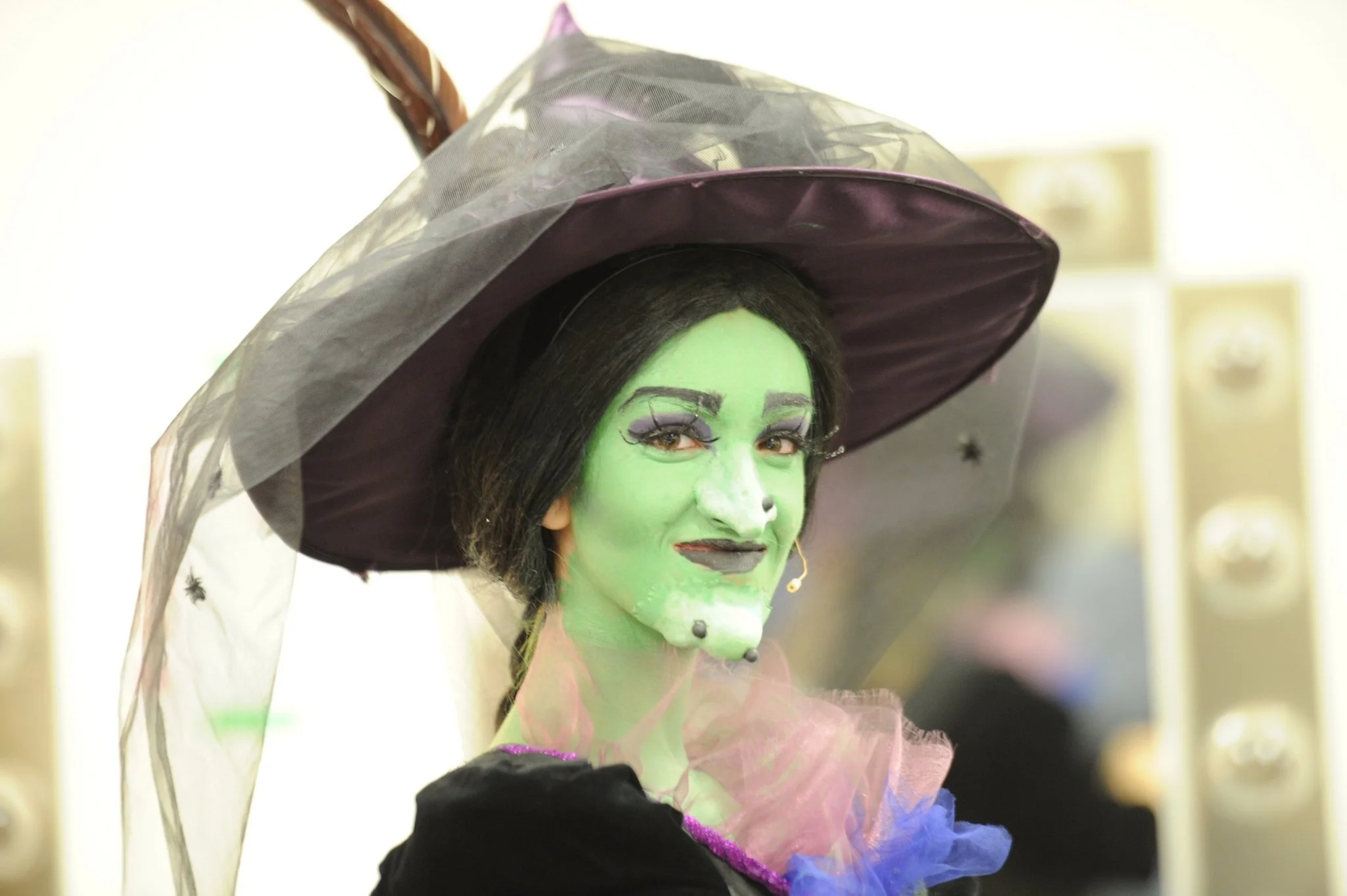 A person dressed as a witch with green face paint, black hair, and wearing a dark purple witch hat with a veil, smiling.