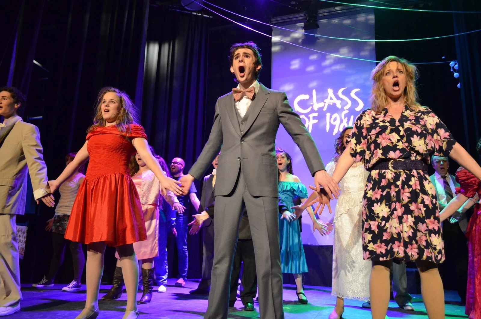 The cast of the musical Grease performing on stage, dressed in colorful costumes, holding hands, and singing, with a backdrop displaying 'Class of 19' and stage lights overhead.