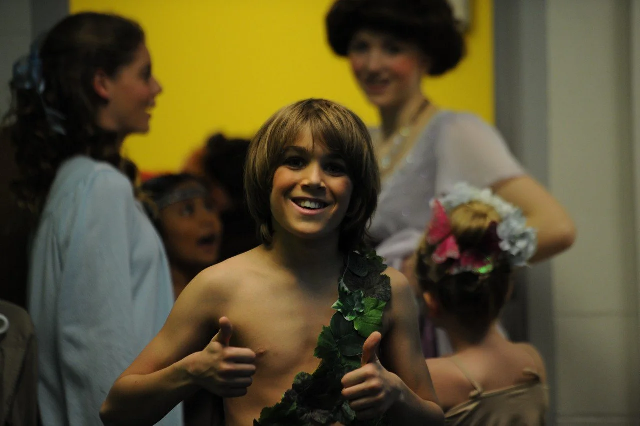 A group of people at a costume party, with a shirtless young man in the foreground wearing a leaf costume giving a thumbs up and smiling.