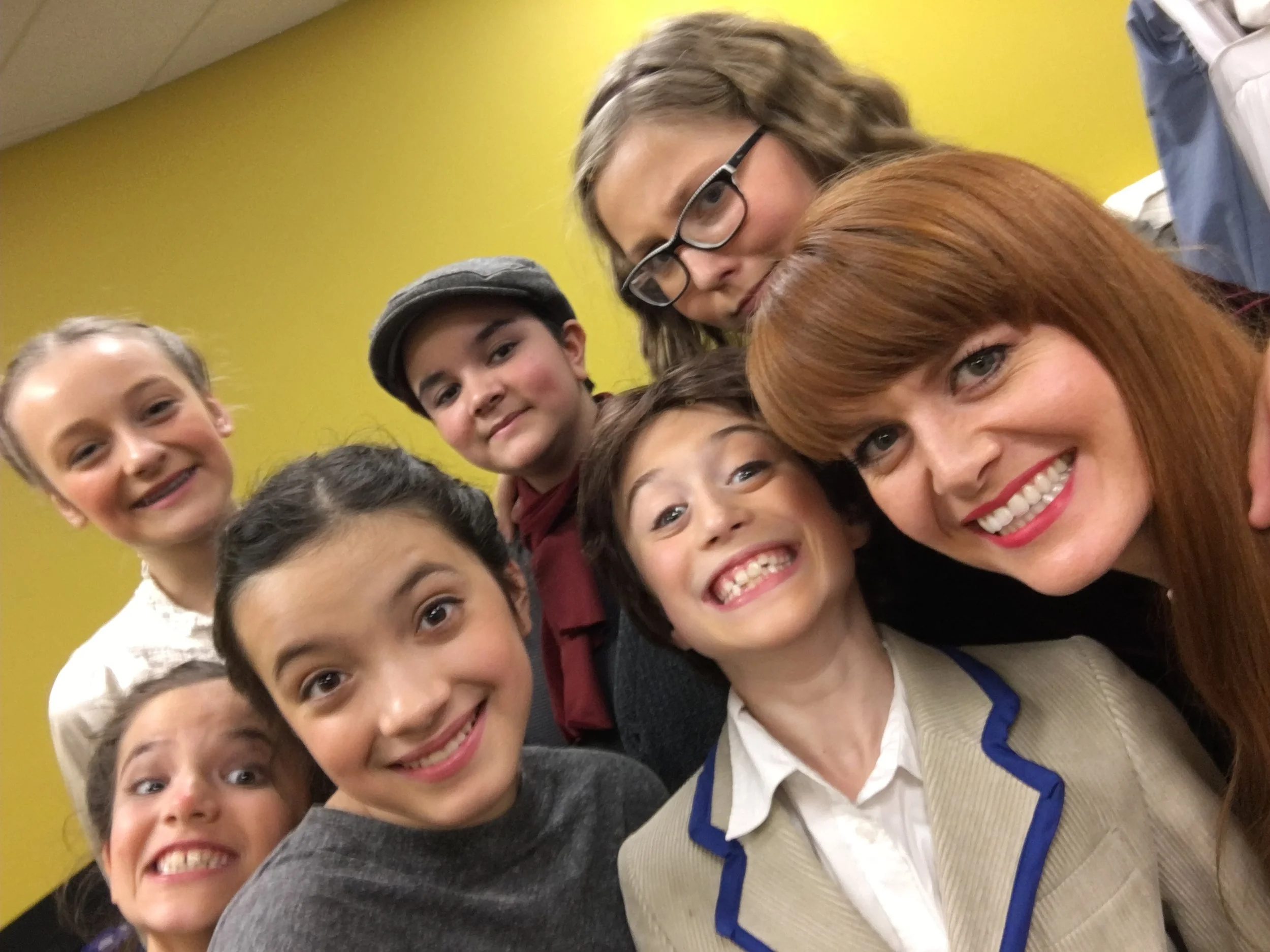A group of children and a woman gathered closely together, smiling for a group photo against a yellow wall.