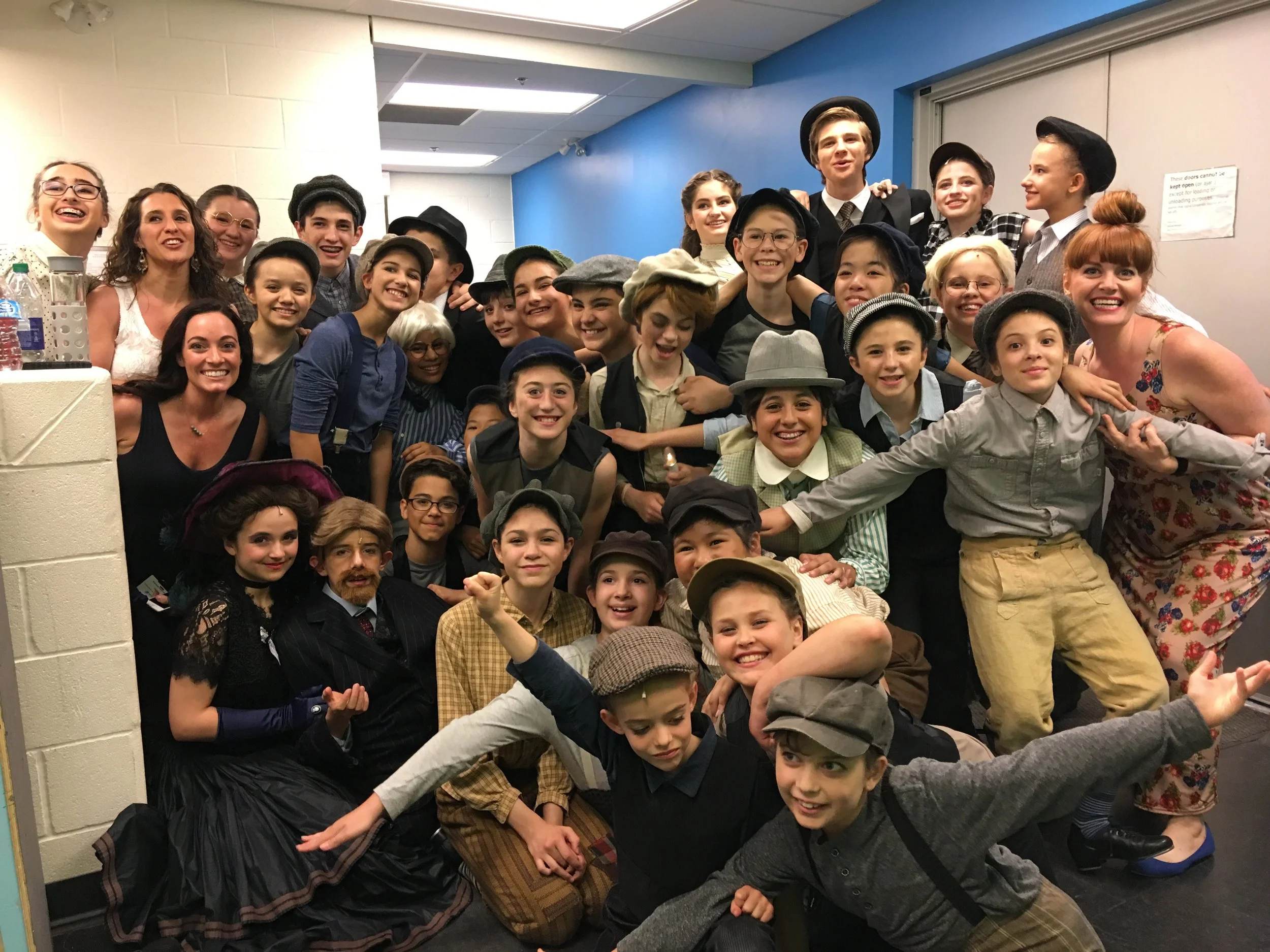 A large group of children and a few adults dressed in vintage clothing, including hats and suspenders, are gathered together in a room, smiling and posing for a photo.