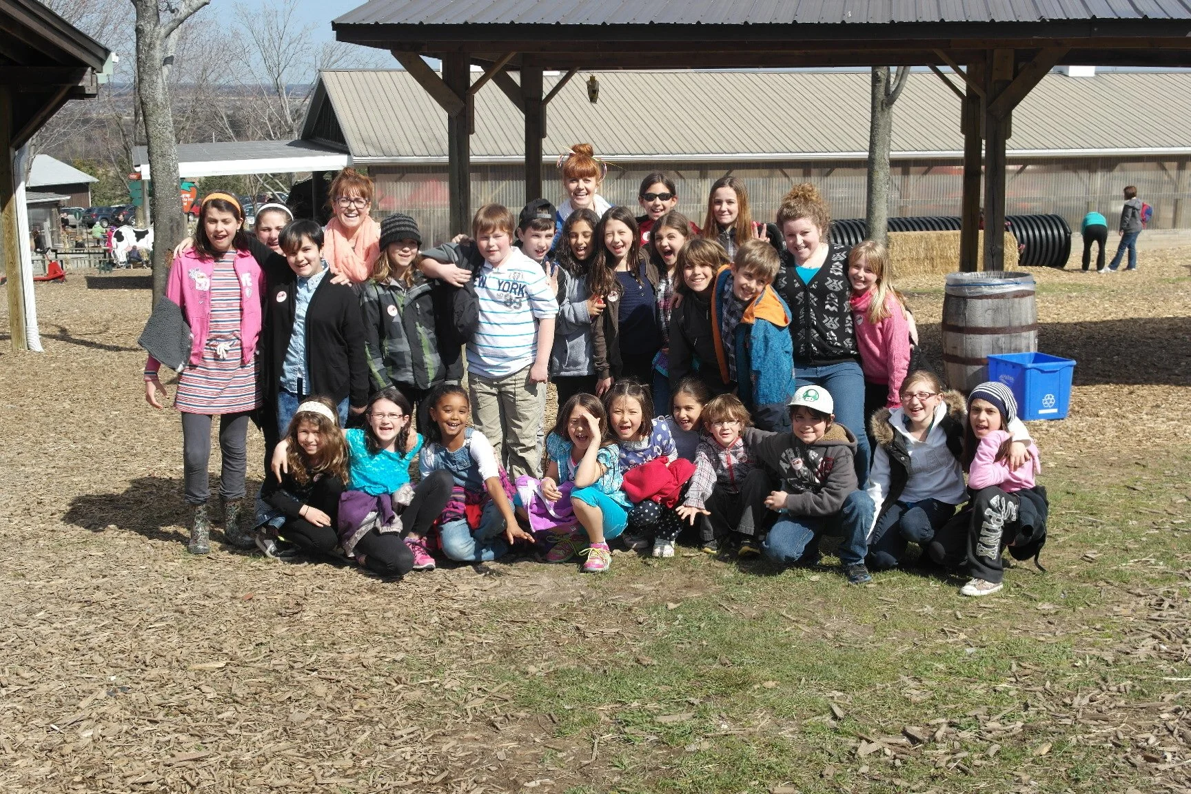 Group of children and a few adults posing outdoors in a wooded area with barns and trees in the background. The children are smiling and sitting or standing together, dressed in casual clothes.