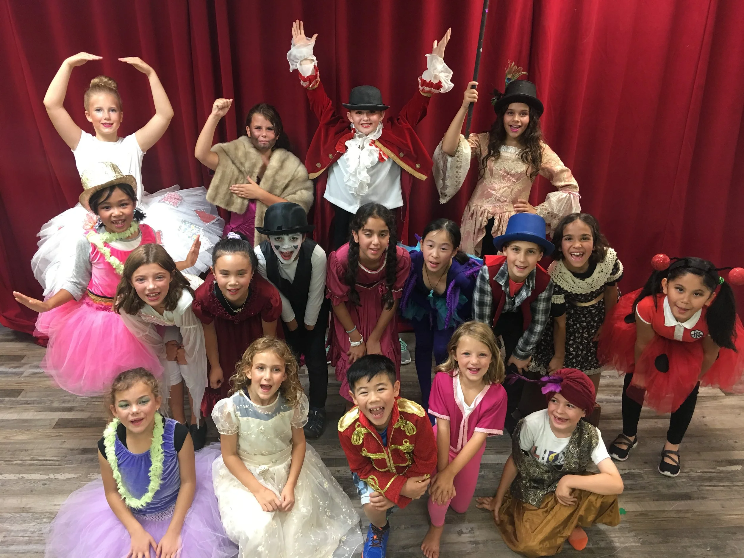 Group of children on stage, dressed in costumes, posing with some raising their arms and others smiling, with a red curtain in the background.