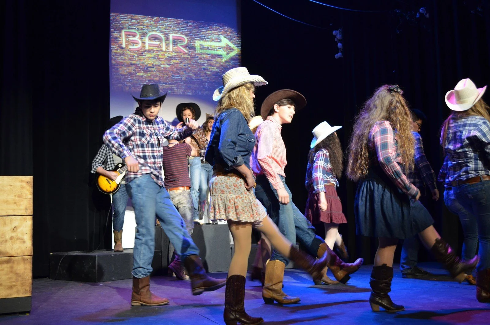 Young people dressed in cowboy attire dancing on stage during a performance, with a sign indicating a bar in the background.