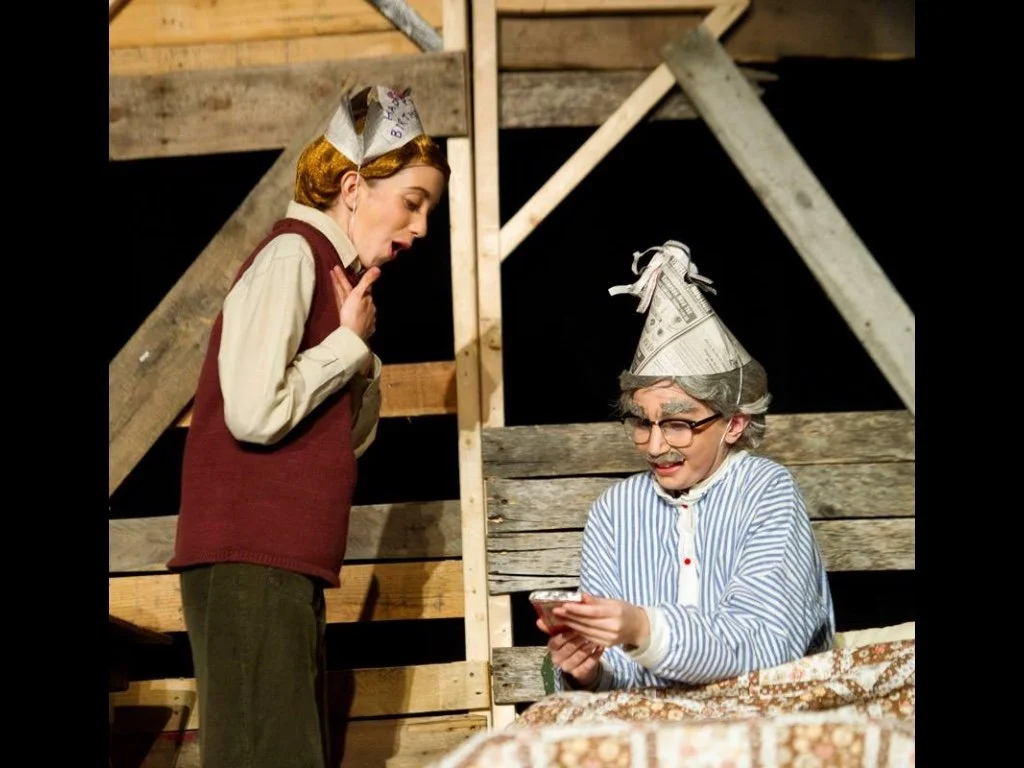 Two actors on stage, one dressed as an elderly woman with gray hair and glasses, wearing a blue and white striped nightgown and a newspaper hat, sitting on a bed and looking at a phone. The other dressed as a younger woman with short brown hair, wear
