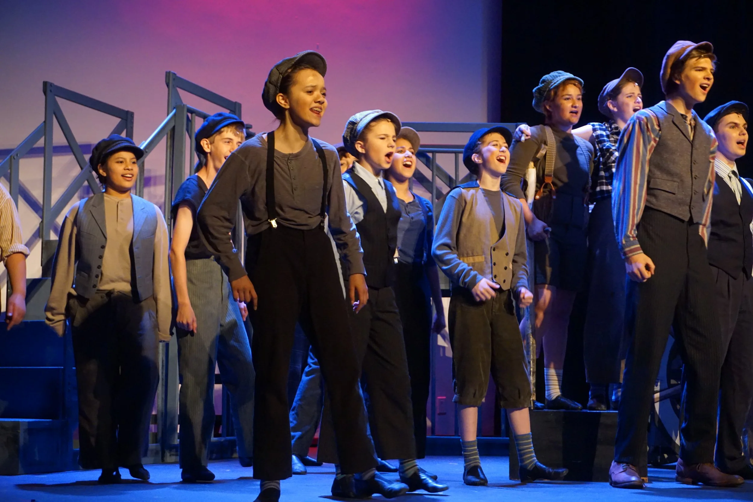 Children dressed in vintage clothing performing on stage with some wearing hats. They are standing in a line, singing or acting out a scene. Stage set includes a structure resembling stairs or a platform.