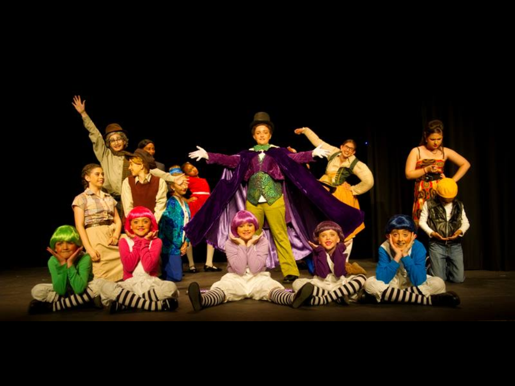 Children dressed in colorful costumes perform on a stage, with a boy in the center wearing a purple cape and top hat as the main character.