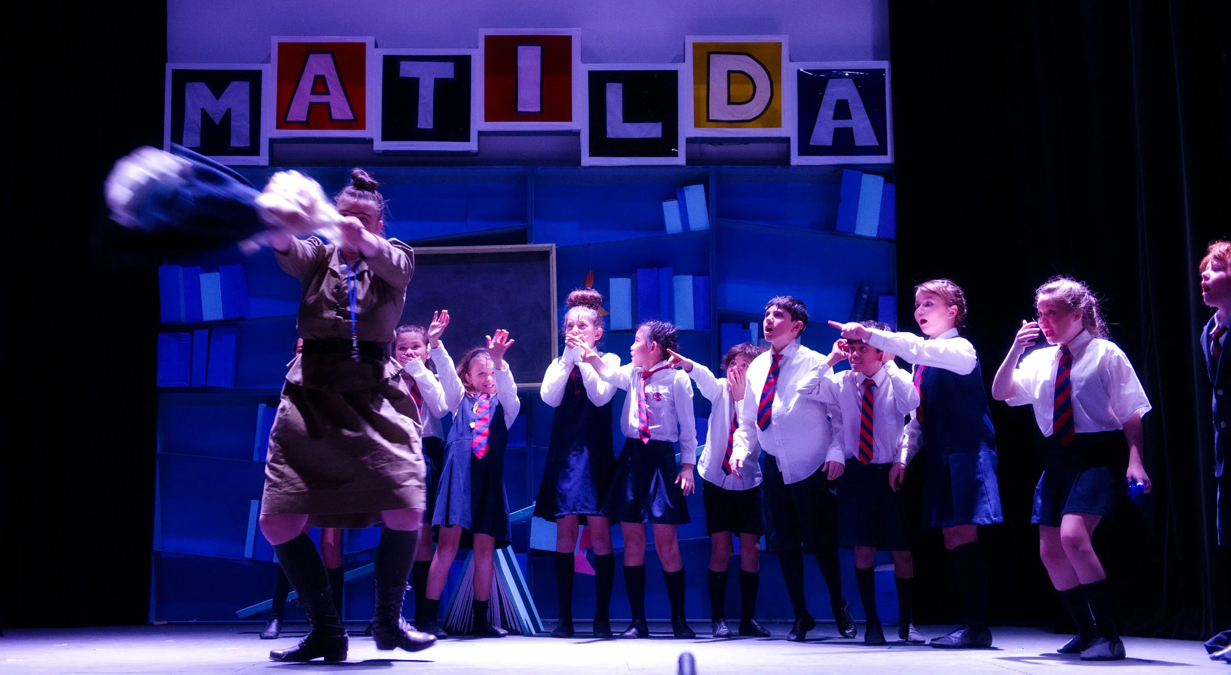 Children in school uniforms on stage during a performance, with a woman in a scout uniform in the foreground and a colorful sign reading 'Matilda' above them.