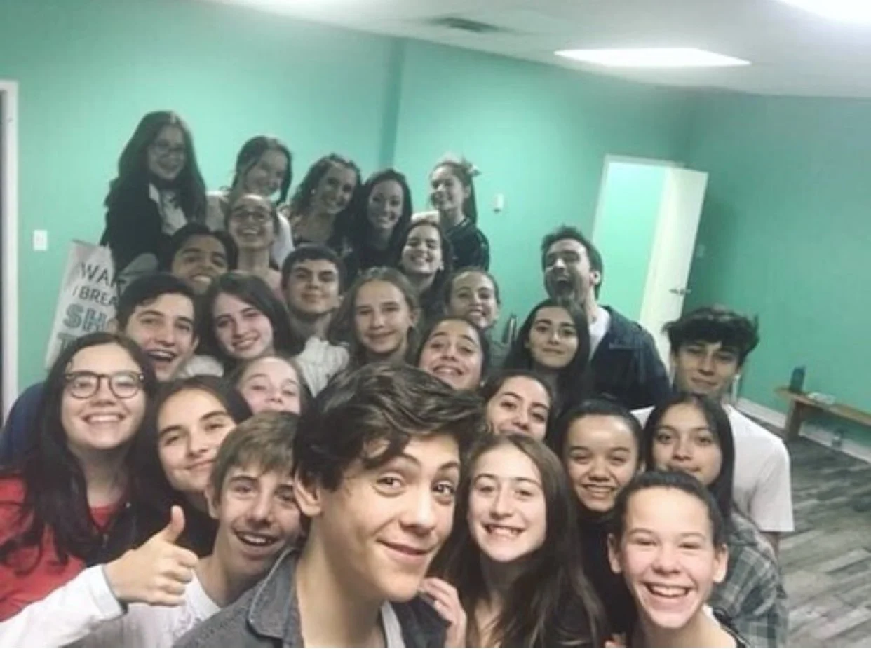 Group of young people smiling and taking a group selfie indoors with green walls.