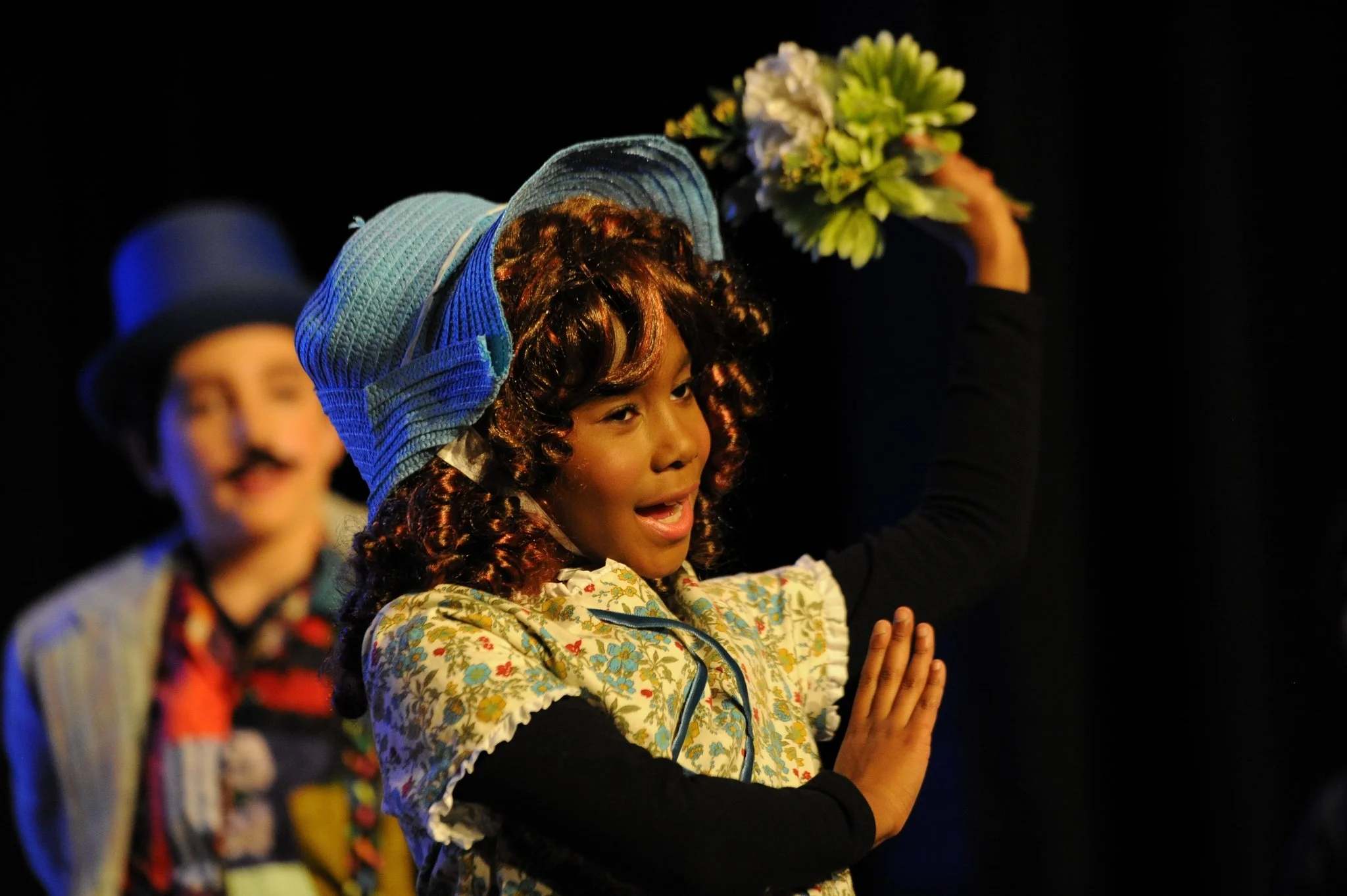 A child with curly hair dressed in a floral costume, holding their hands together in prayer, with a person in the background and dark lighting.