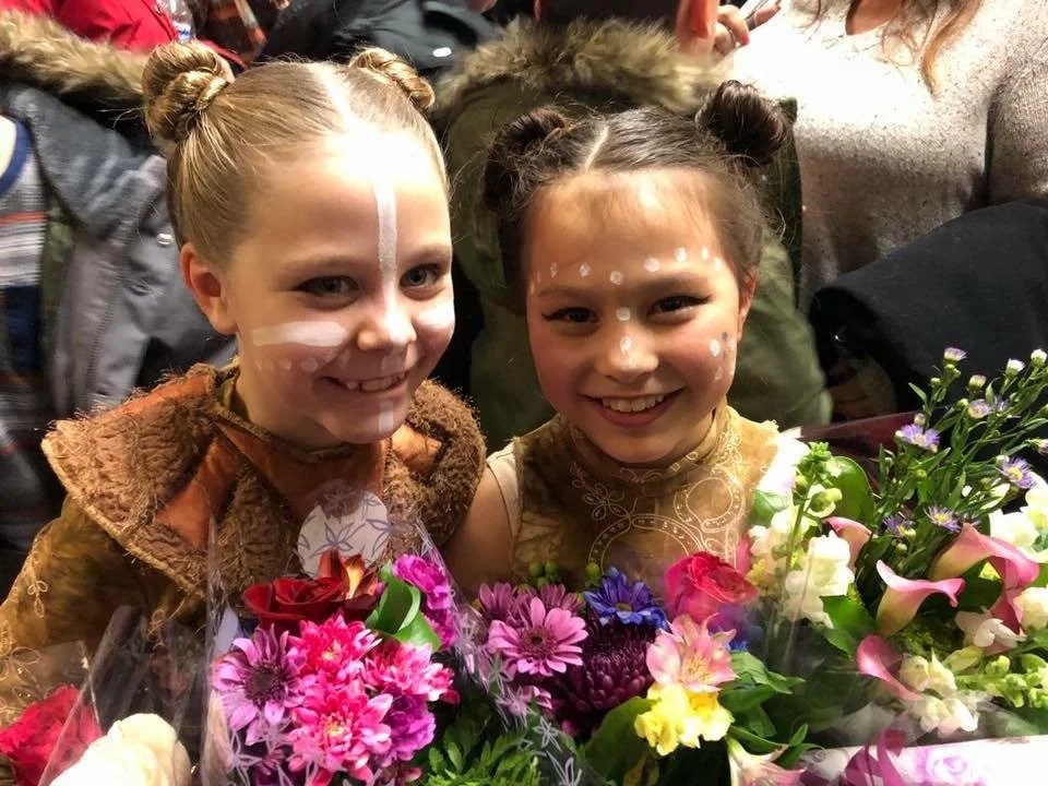 Two young girls smiling and holding colorful flower bouquets, dressed in costumes with face paint, inside a crowded room.