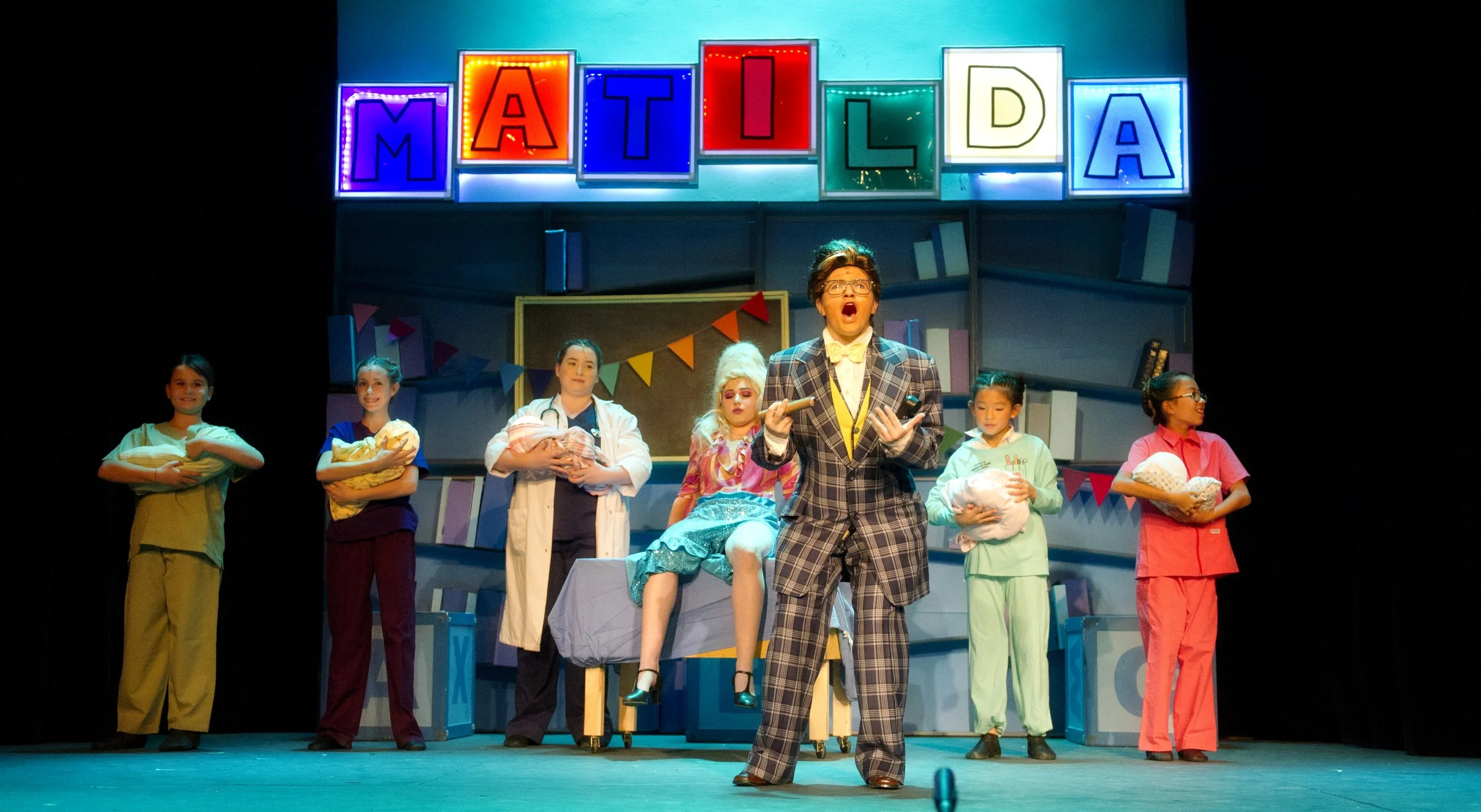 Stage scene from the musical 'Matilda' with actors in costumes. The large colorful sign reads 'MATILDA' in illuminated blocks. A male actor in a checkered suit and glasses stands at center, surrounded by children holding pillows, with a woman in a wh