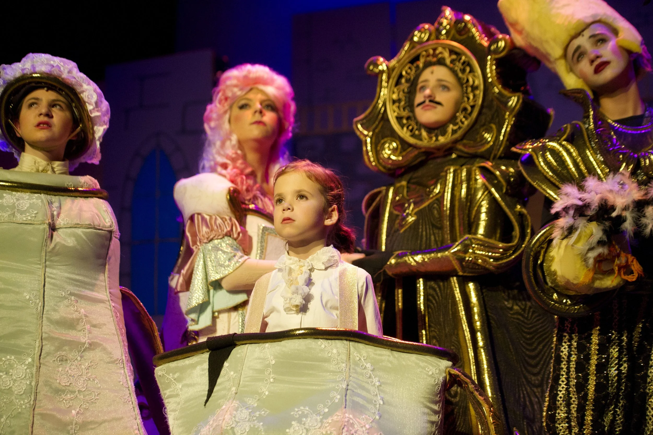 Children dressed as characters in a theatrical performance, with costumes that evoke historical or fantasy themes, including large hats and ornate outfits, standing around a large, decorated drum.