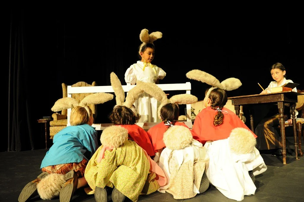 Children participating in a play with actors dressed as rabbits and mice, sitting on stage with props and a table.