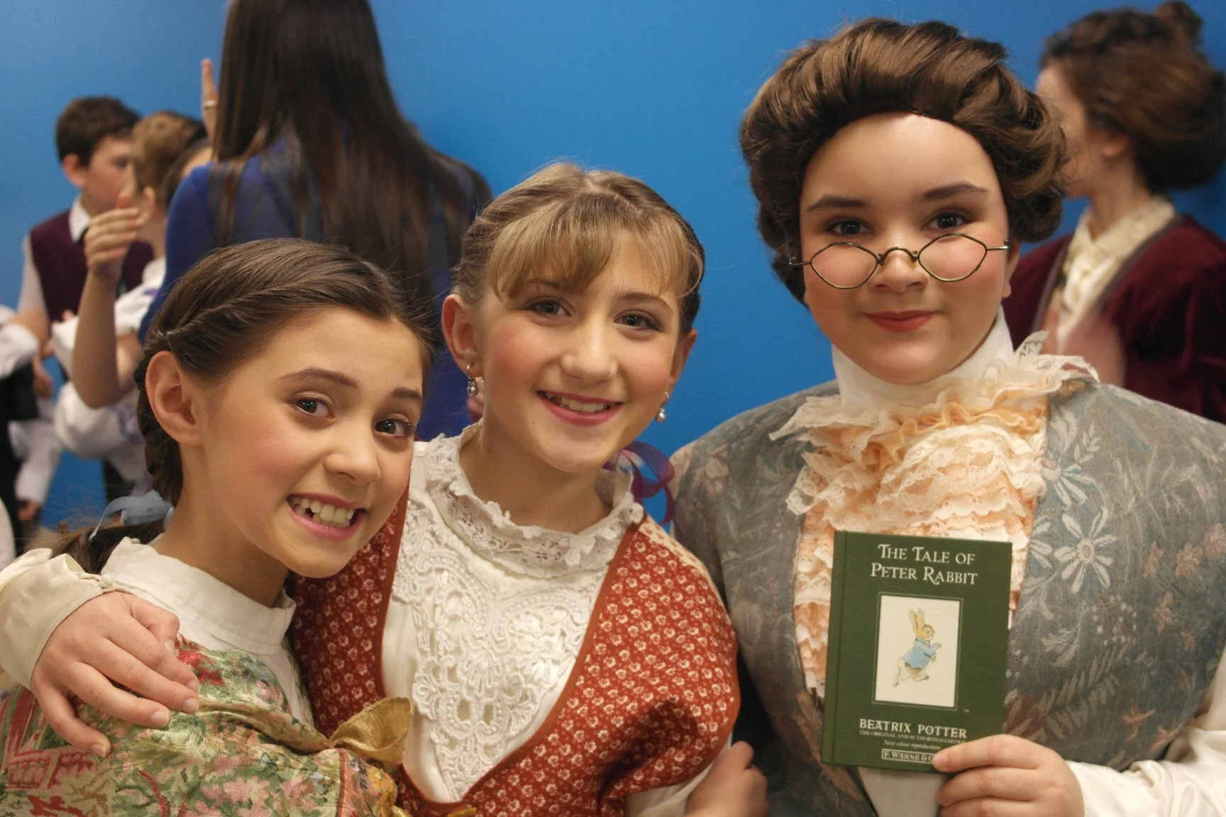 Three girls in vintage clothing at a book event, with one holding a copy of 'The Tale of Peter Rabbit' by Beatrix Potter.
